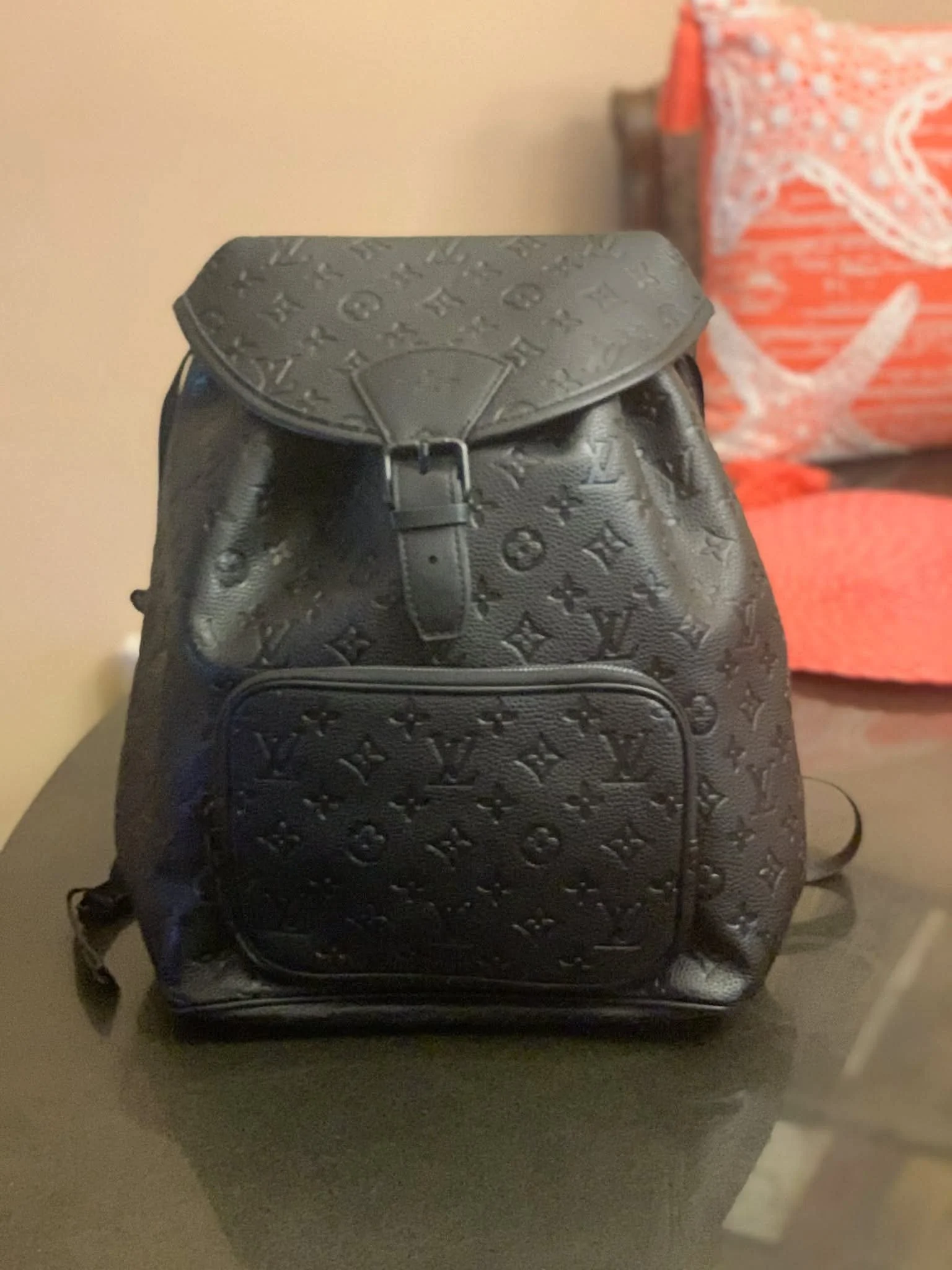 A black Louis Vuitton backpack with the classic monogram pattern, placed on a table in front of a beige wall and a red patterned bag in the background.
