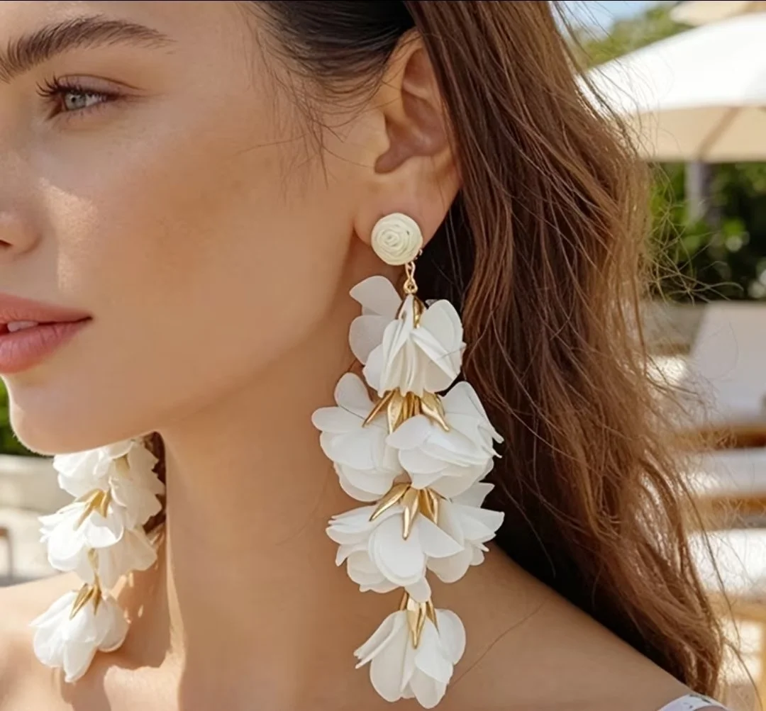 Close-up of a woman wearing large white floral earrings with gold accents, outdoors with a background of greenery and a white umbrella.