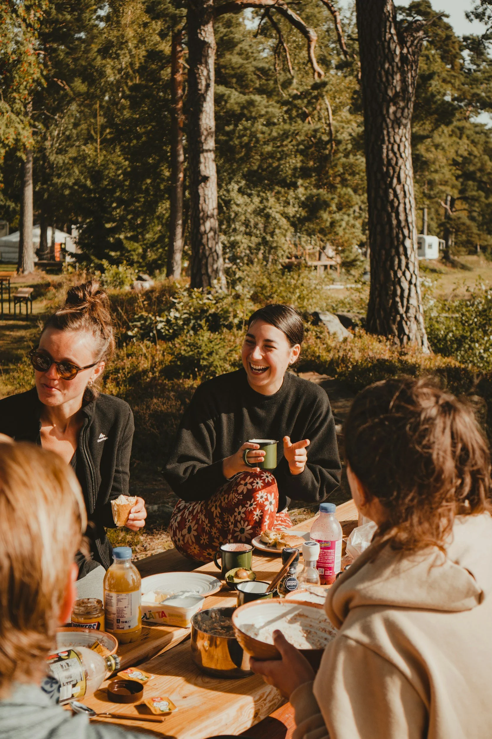 People sitting outside at a picnic table enjoying food and drinks, surrounded by tall trees and greenery on a sunny day.