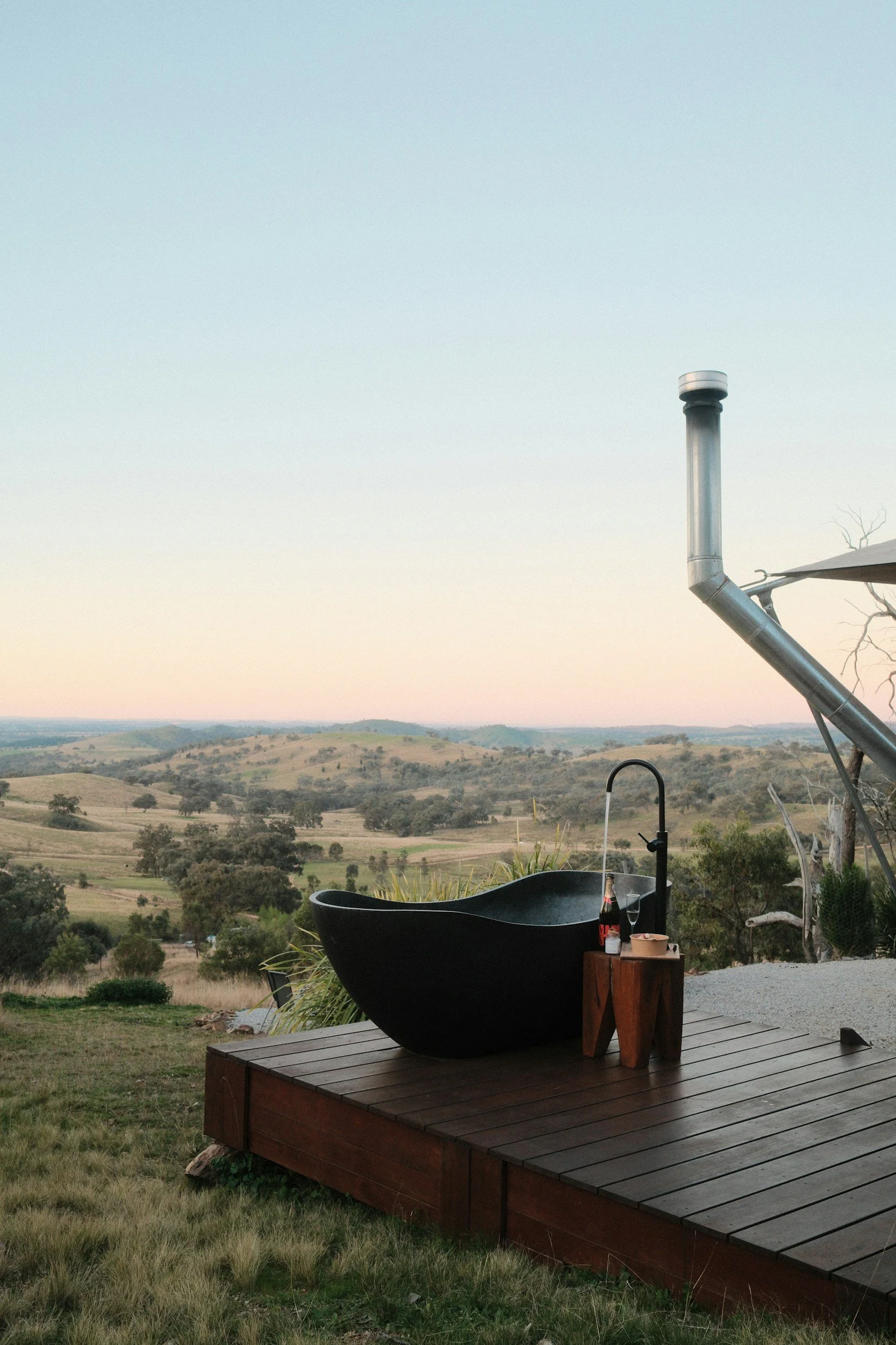 A large black outdoor bathtub on a wooden deck overlooking rolling hills and a green landscape with trees, under a clear sky at sunset.