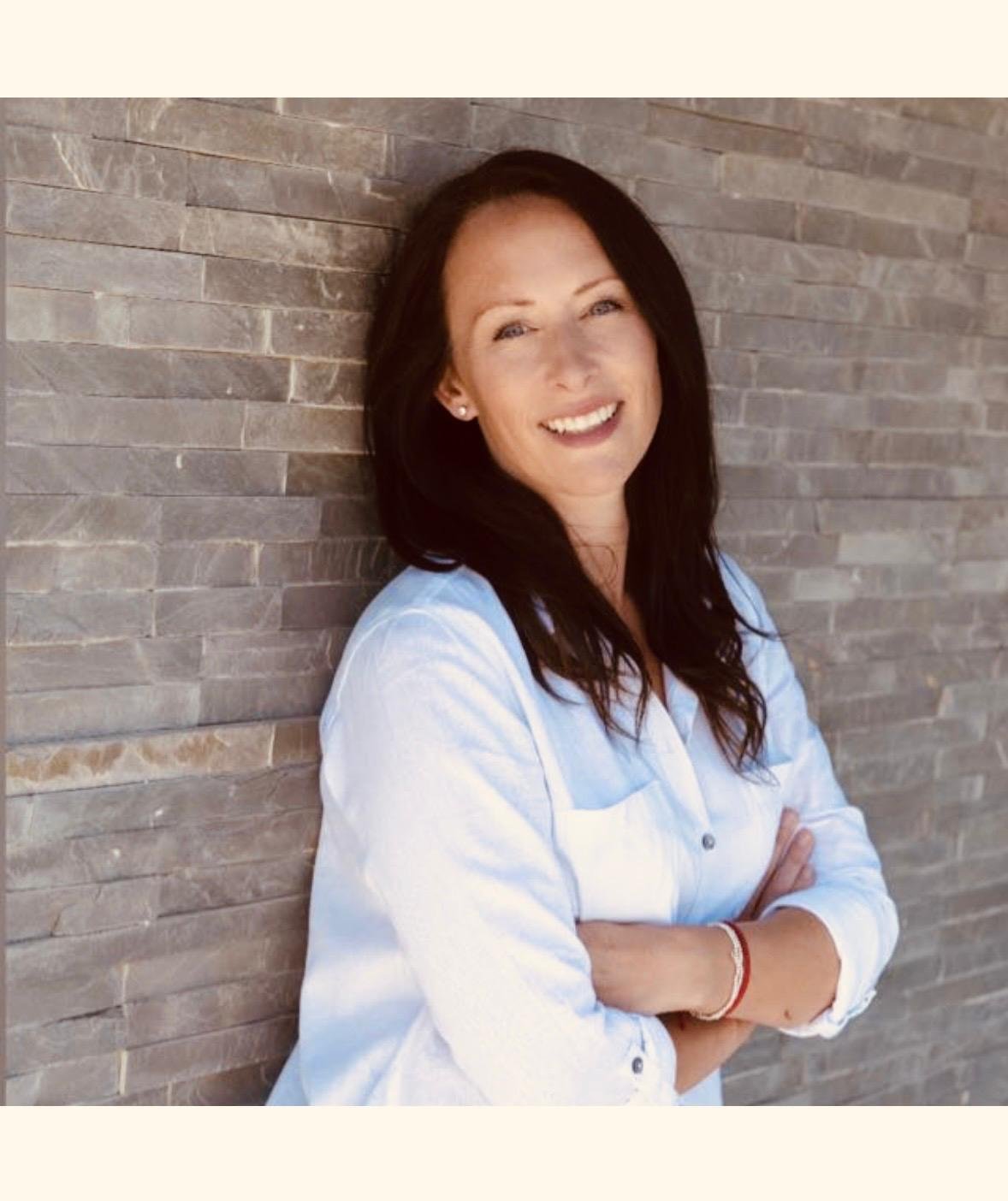 A woman with long dark hair smiling, standing with arms crossed in front of a brick wall, wearing a light-colored shirt.