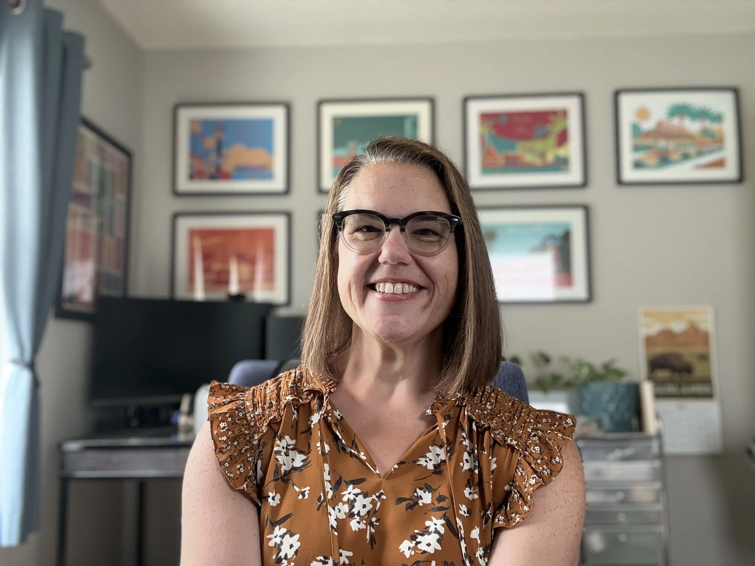 A woman with long brown hair and glasses smiling, wearing a brown floral top, sitting in a room with framed colorful artwork on the wall behind her.