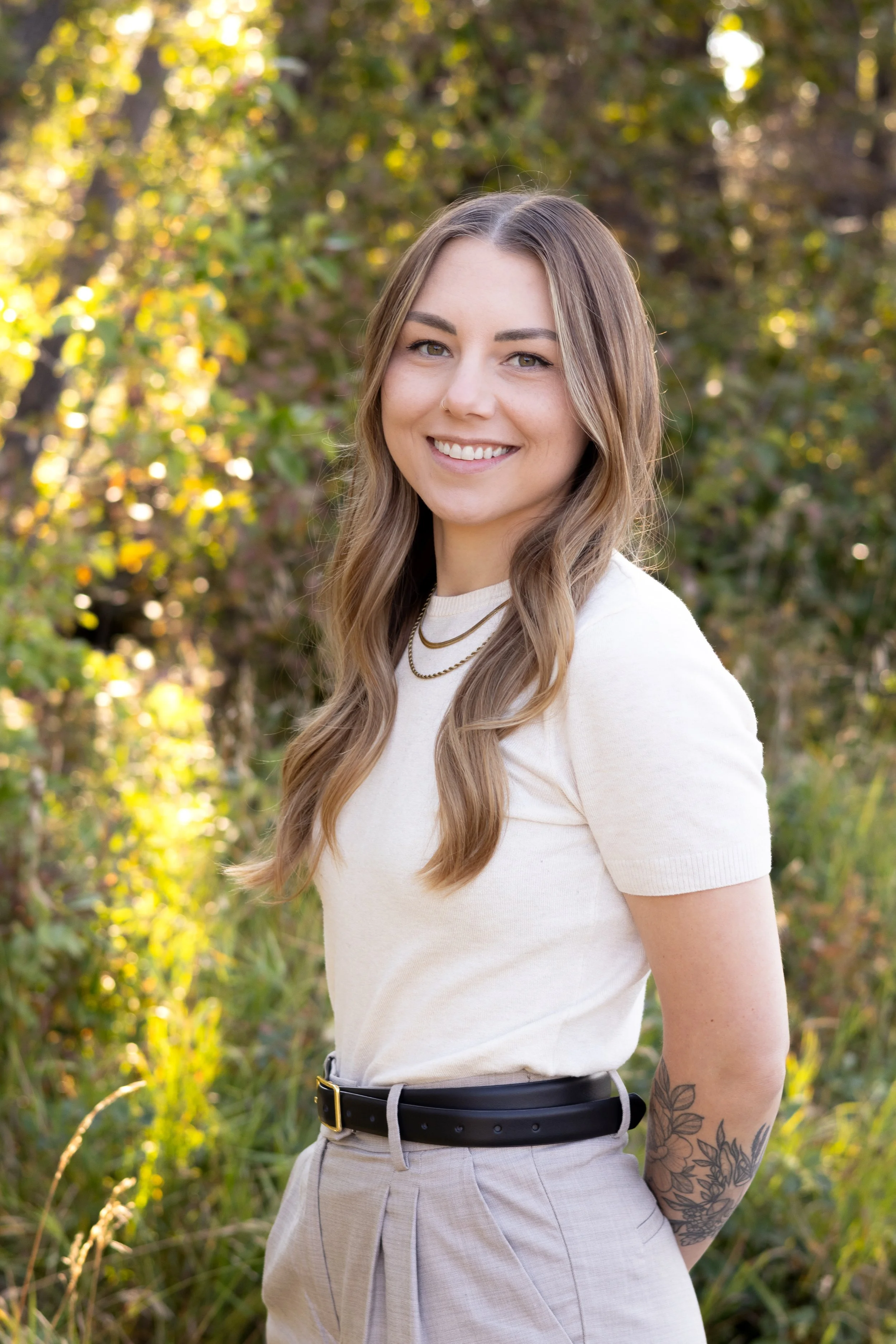 A young woman with long, wavy, light brown hair and a nose ring, smiling outdoors in a natural setting with green and yellow foliage in the background. She is wearing a white short-sleeve shirt, layered necklaces, beige high-waisted pants with a black belt, and has floral tattoos on her left forearm.