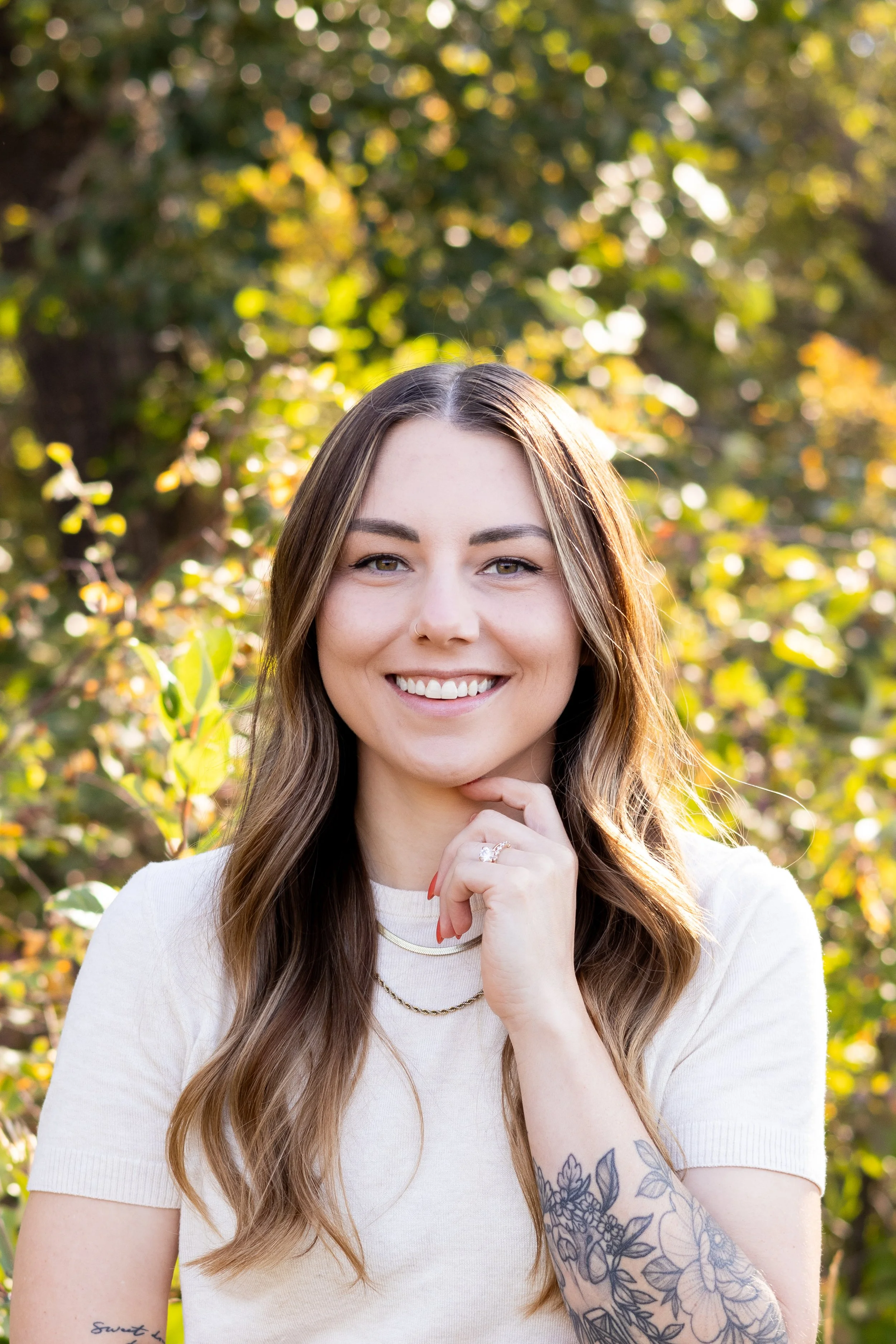 A young woman with long, wavy brown hair and piercing green eyes smiles outdoors in front of a background of blurred green and yellow foliage. She is wearing a white shirt and has tattoos on her left forearm and right wrist. Her right hand is lightly touching her chin, and she appears happy and relaxed.