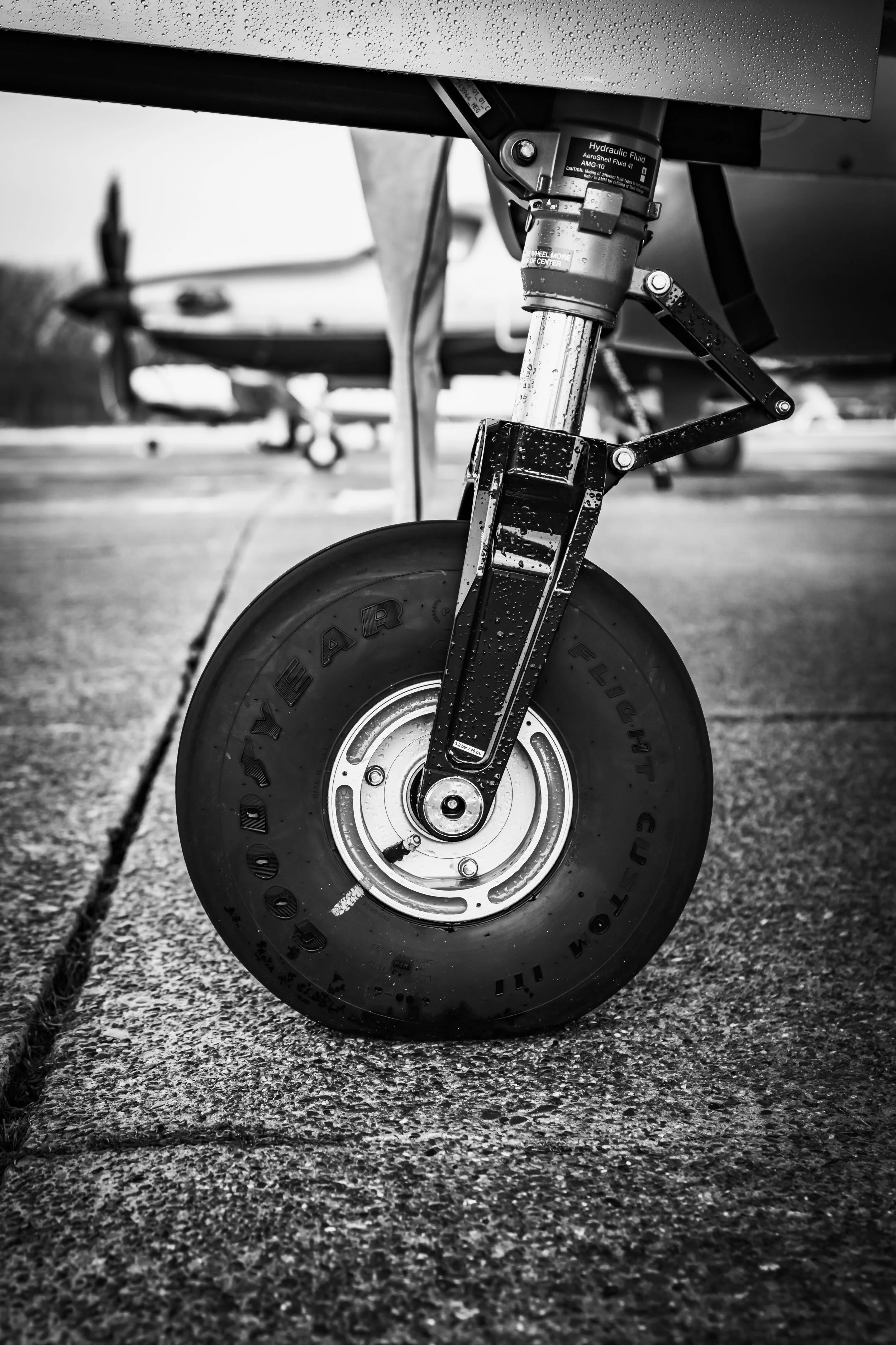 Close-up black and white photo of an aircraft nose wheel with a blurred airplane in the background, wet surface, and raindrops on the aircraft part.