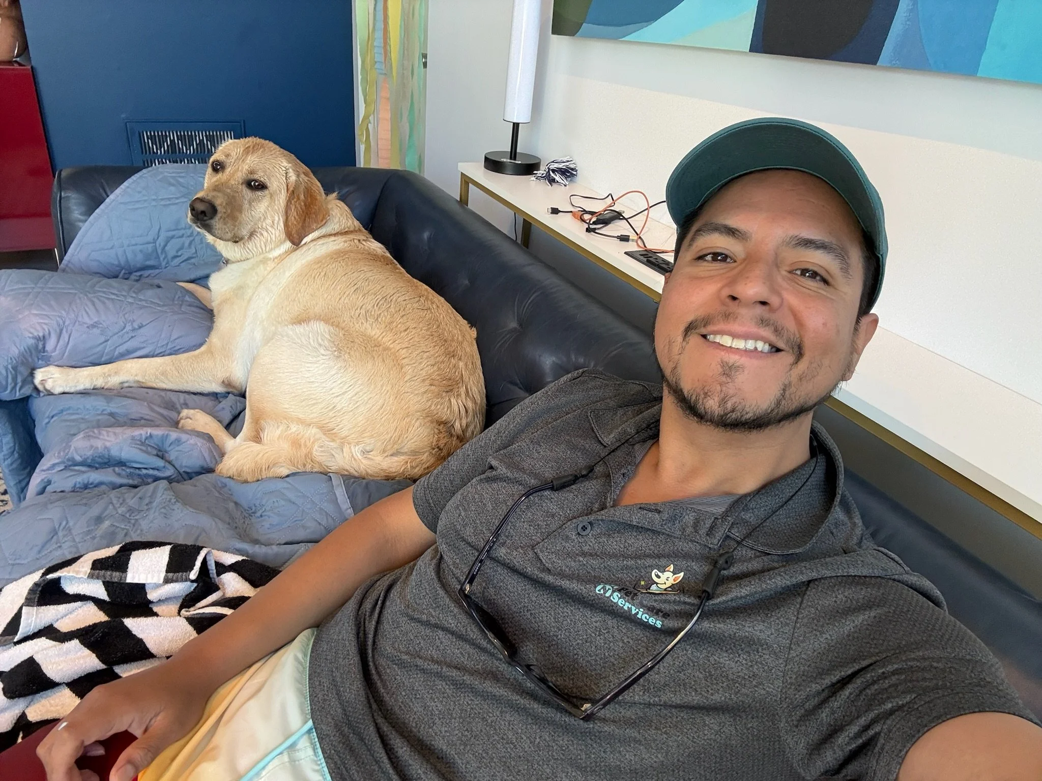 A man taking a selfie with a yellow Labrador Retriever dog laying on a black couch next to him in a room with colorful wall art, a white side table with a lamp, cords, and a black-and-white striped blanket.