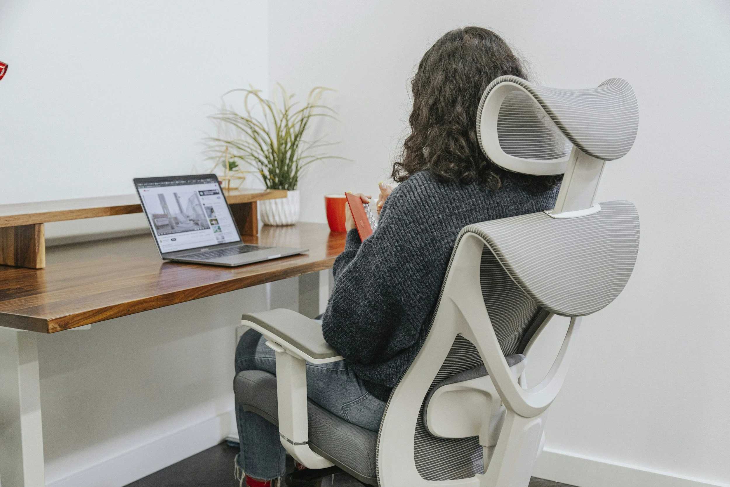 Person with curly hair sitting in an ergonomic office chair at a wooden desk, looking at a tablet and with a laptop open in front of them, decorated with a plant and a red cup.