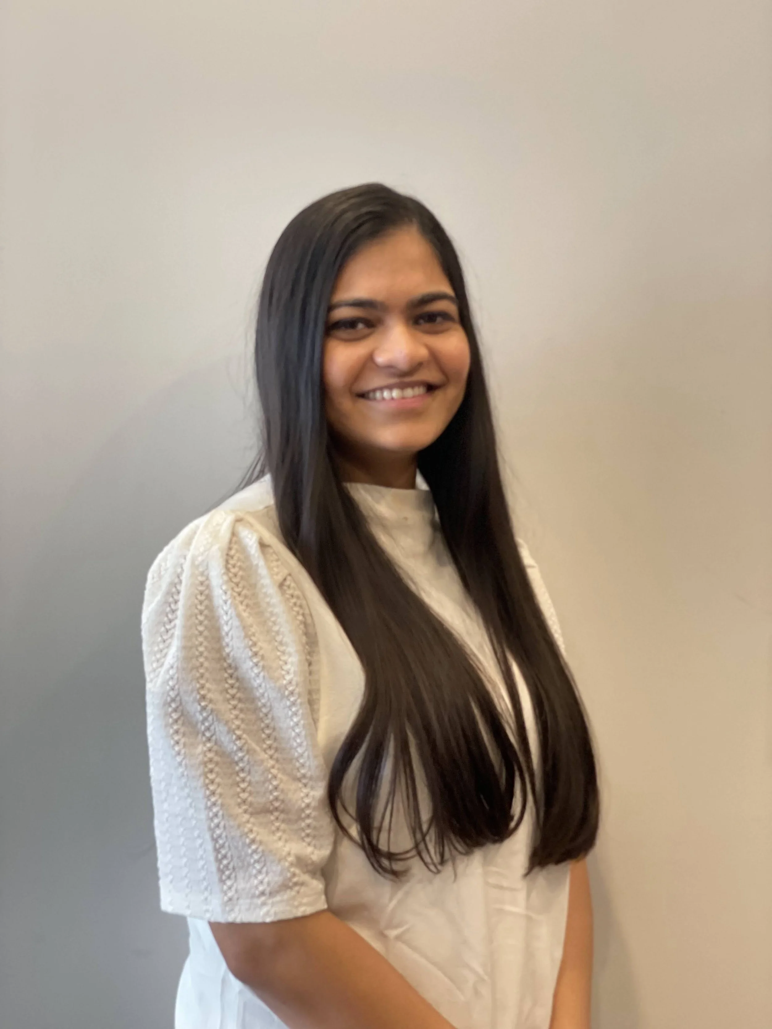 A woman with long dark hair smiling, standing against a plain light gray wall, wearing a cream-colored knitted sweater.