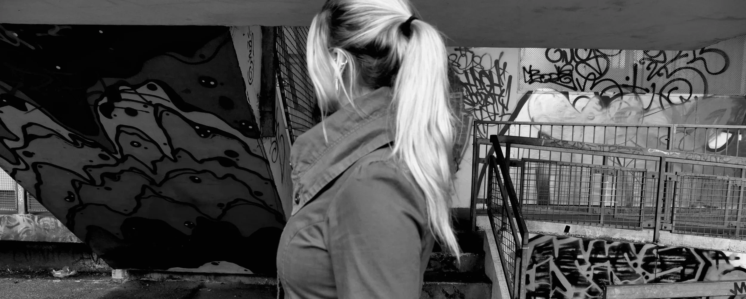 A woman with long blonde hair in a ponytail is standing in front of a graffiti-covered wall at a skatepark.