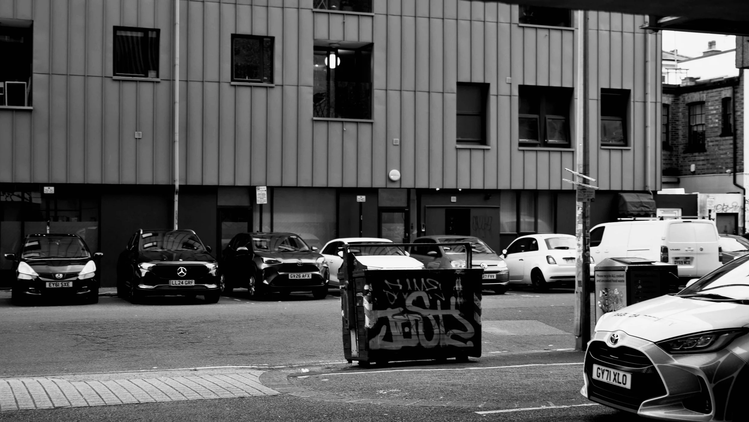 A black and white photo of a city street scene with parked cars in front of a modern building with vertical siding and multiple windows.