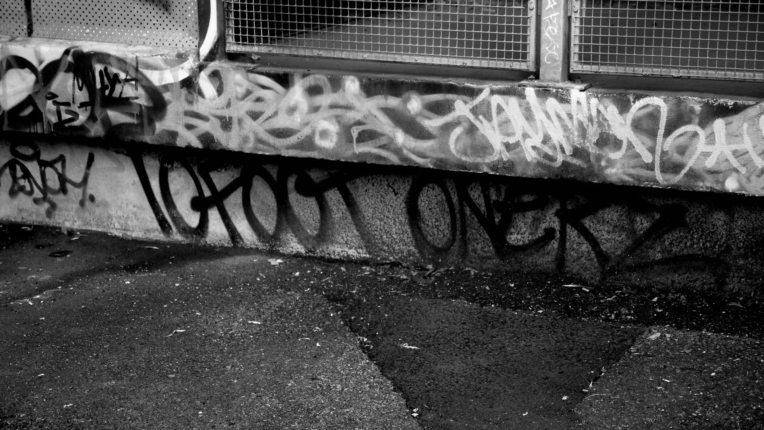 Black and white photo of a concrete wall with graffiti, metal mesh grid, and a ground covered in dirt and small debris.