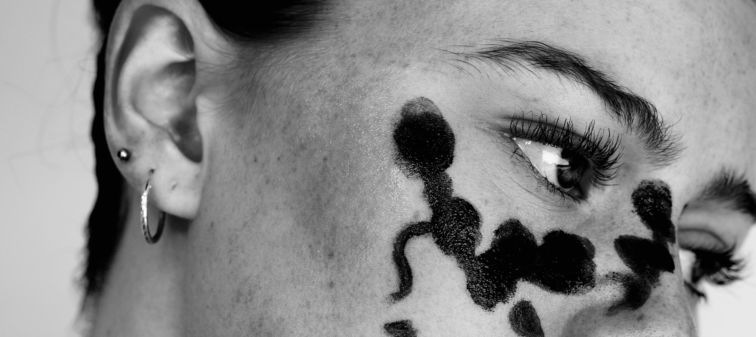Close-up of a person's face with bold black face paint and tattoos, focusing on the eye, eyebrow, and ear, with tattoos on the cheek and a small hoop earring.
