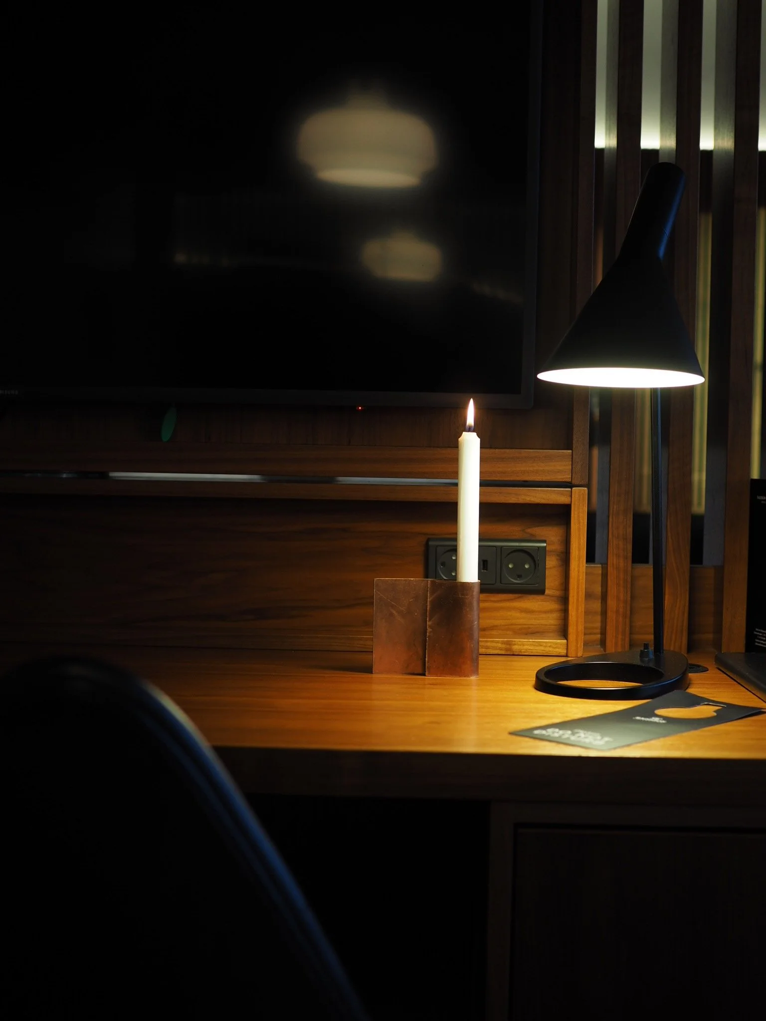 Et rom med mørkt treverk, der en lampe og et lysestake med stearinlys står på et skrivebord. Det er en mørk skjerm på veggen og noen telys i bakgrunnen.
