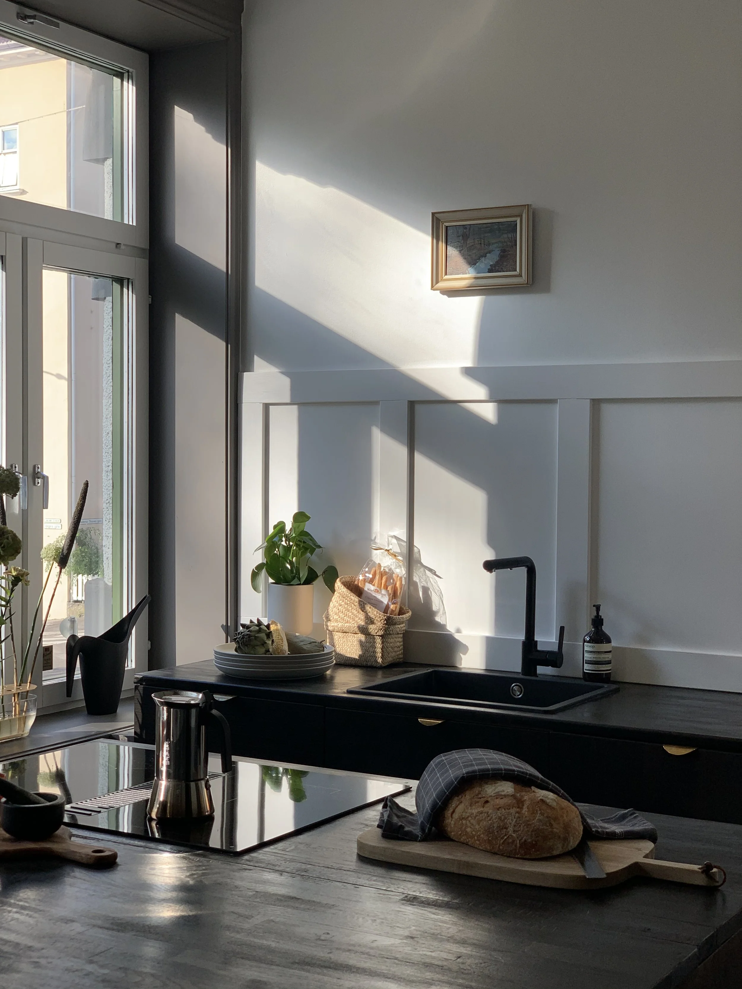 Et kjøkken med svart benkeplate, en svart vask med svart kran, en brød som ligger på et skjærebrett, en kaffetrakter, en plante i hvit potte, og et glass med decor.