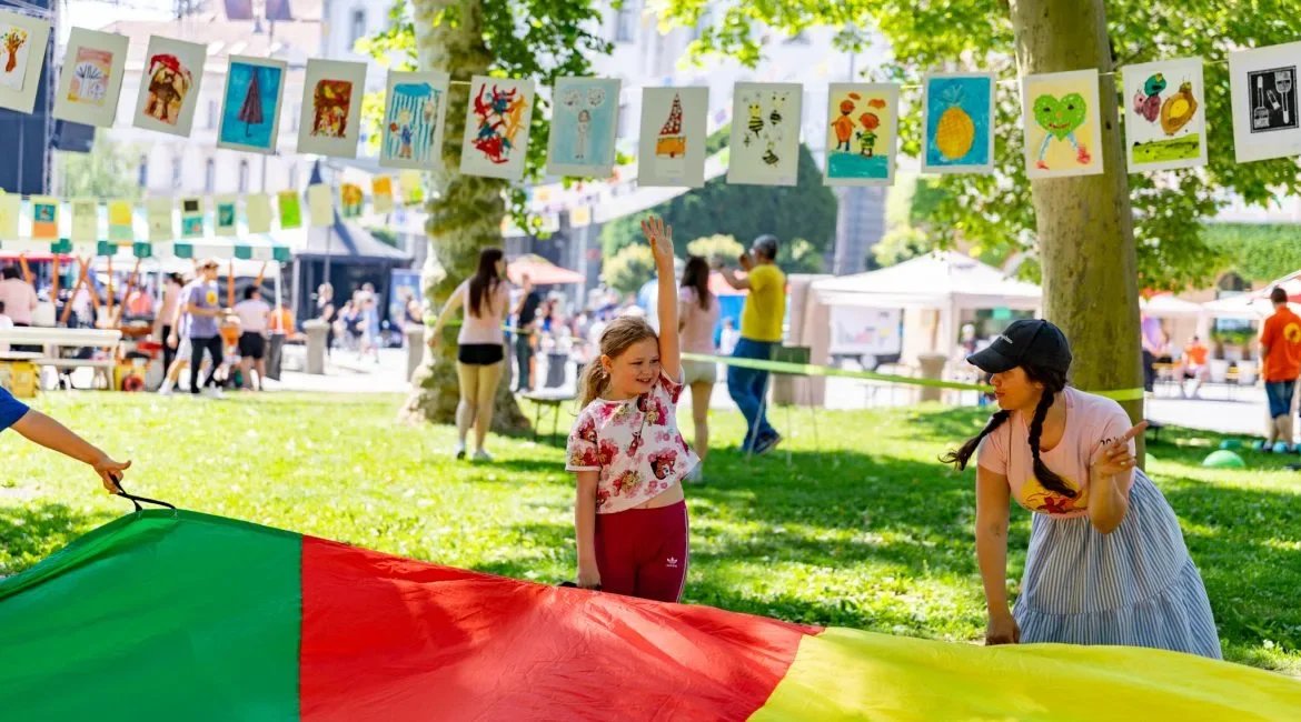 A girl and a woman enjoying a sunny outdoor event with colorful artwork hanging from trees and people in the background.