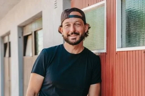 Man with a beard and backwards cap smiling outdoors against a red wooden wall.