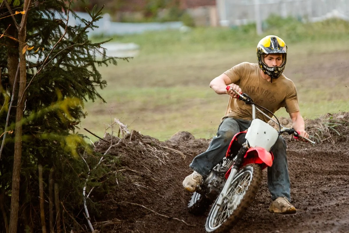 A man wearing a black and yellow helmet and brown t-shirt riding a dirt bike on a dirt trail, leaning into a turn near some bushes.