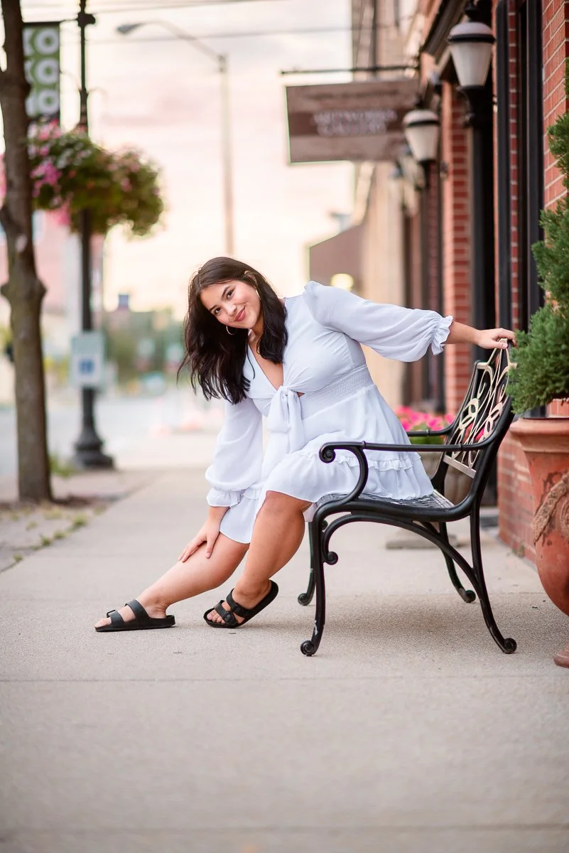 A woman wearing a white dress and black sandals sitting on a black metal park bench on a sidewalk, with her right leg extended outward and her left arm holding the back of the bench. She is smiling at the camera with dark, wavy hair, and there are plants and storefronts in the background.