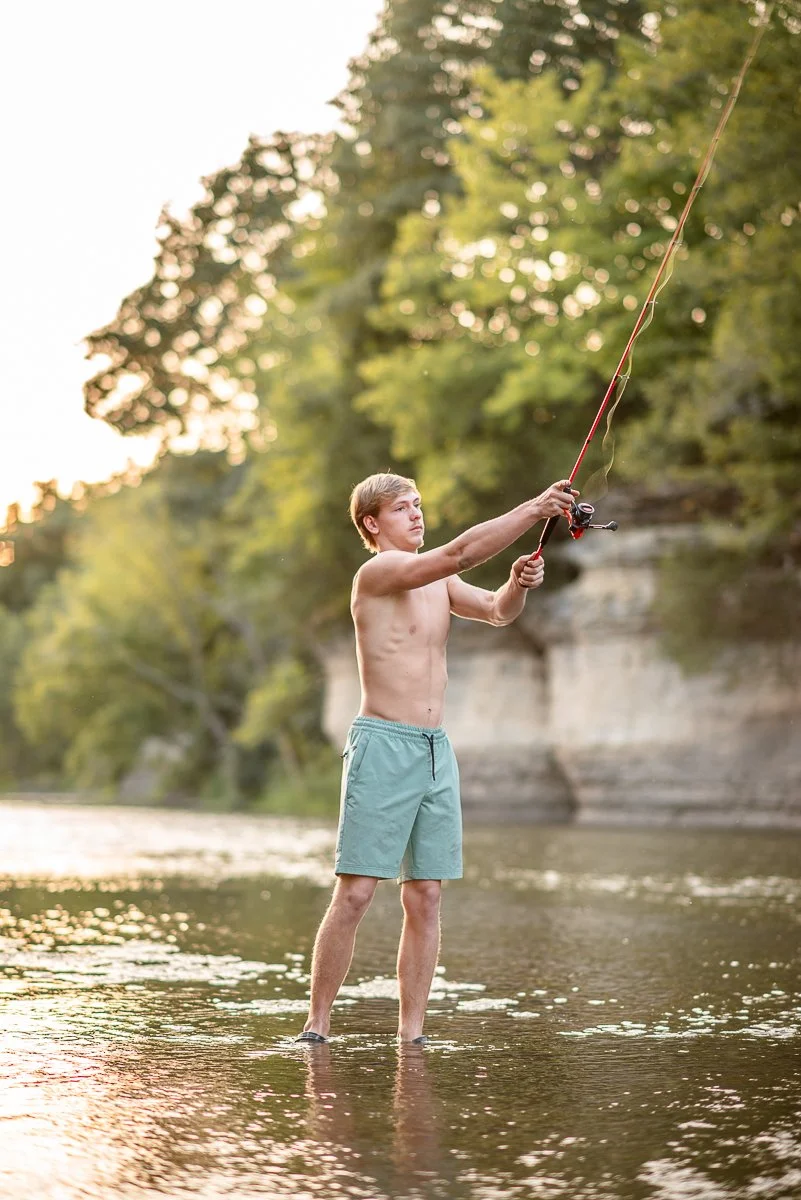 A young man fishing in a river, standing in water up to his ankles, with a forested area and cliffs in the background during sunset.