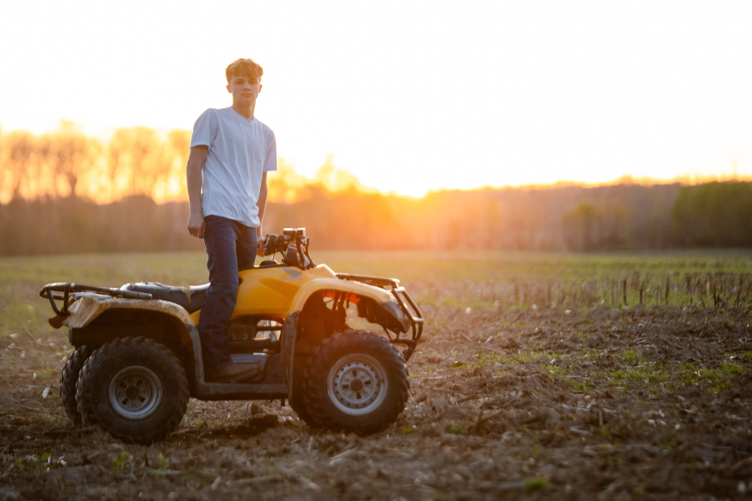 A young person standing on an all-terrain vehicle (ATV) in a field during sunset.