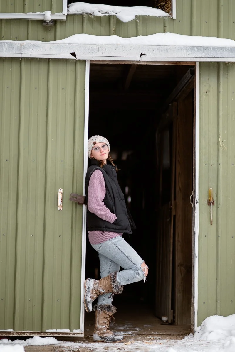 A young woman wearing glasses, a knit hat, a pink sweater, a black vest, ripped jeans, and winter boots, standing in the doorway of a green shed with snow outside.