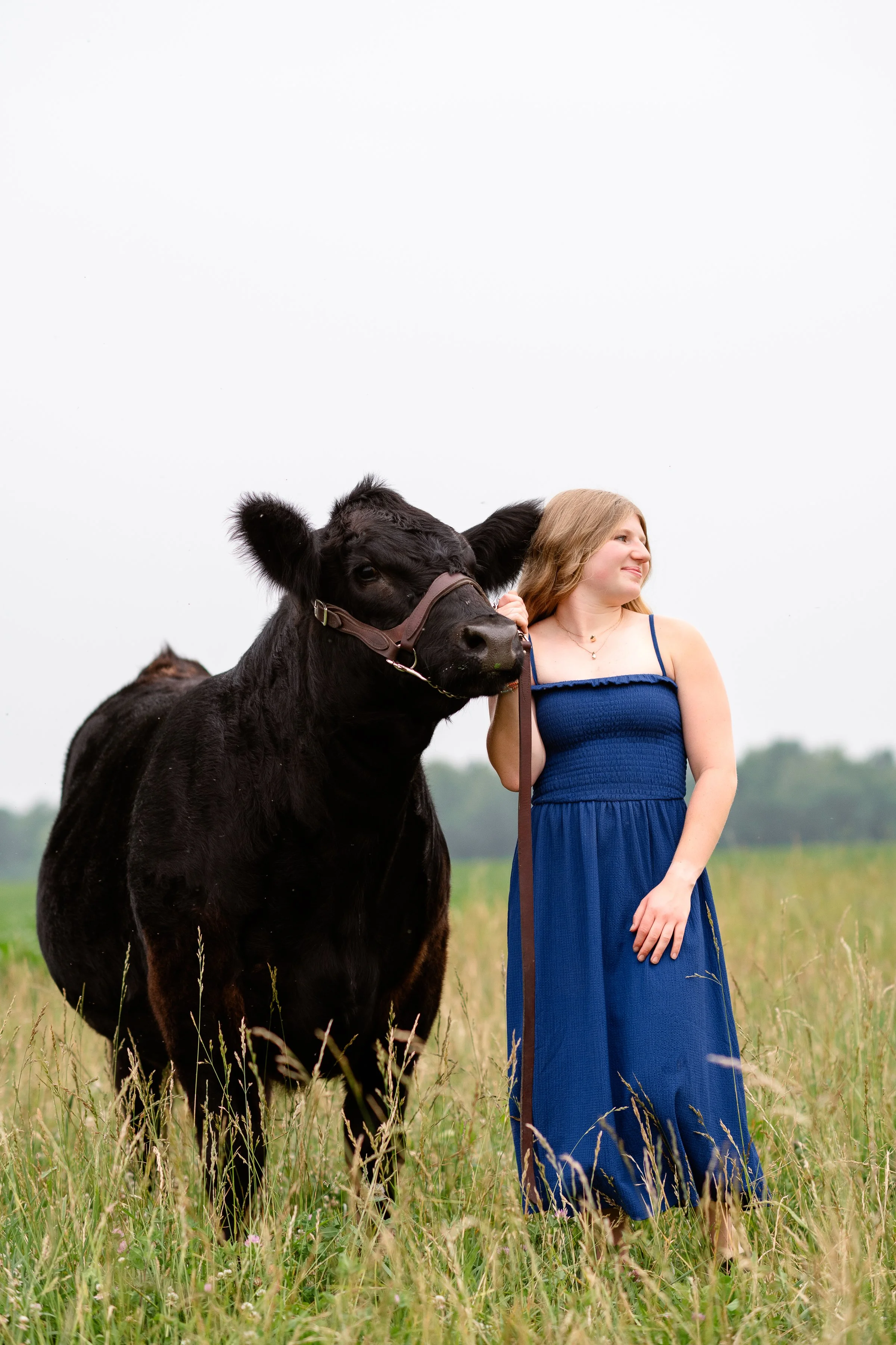 A woman in a blue dress standing in a grassy field, holding a black calf on a leash. The woman is smiling and looking to the right, while the calf is looking forward, wearing a halter. The background is cloudy and the setting appears to be a rural outdoor area.