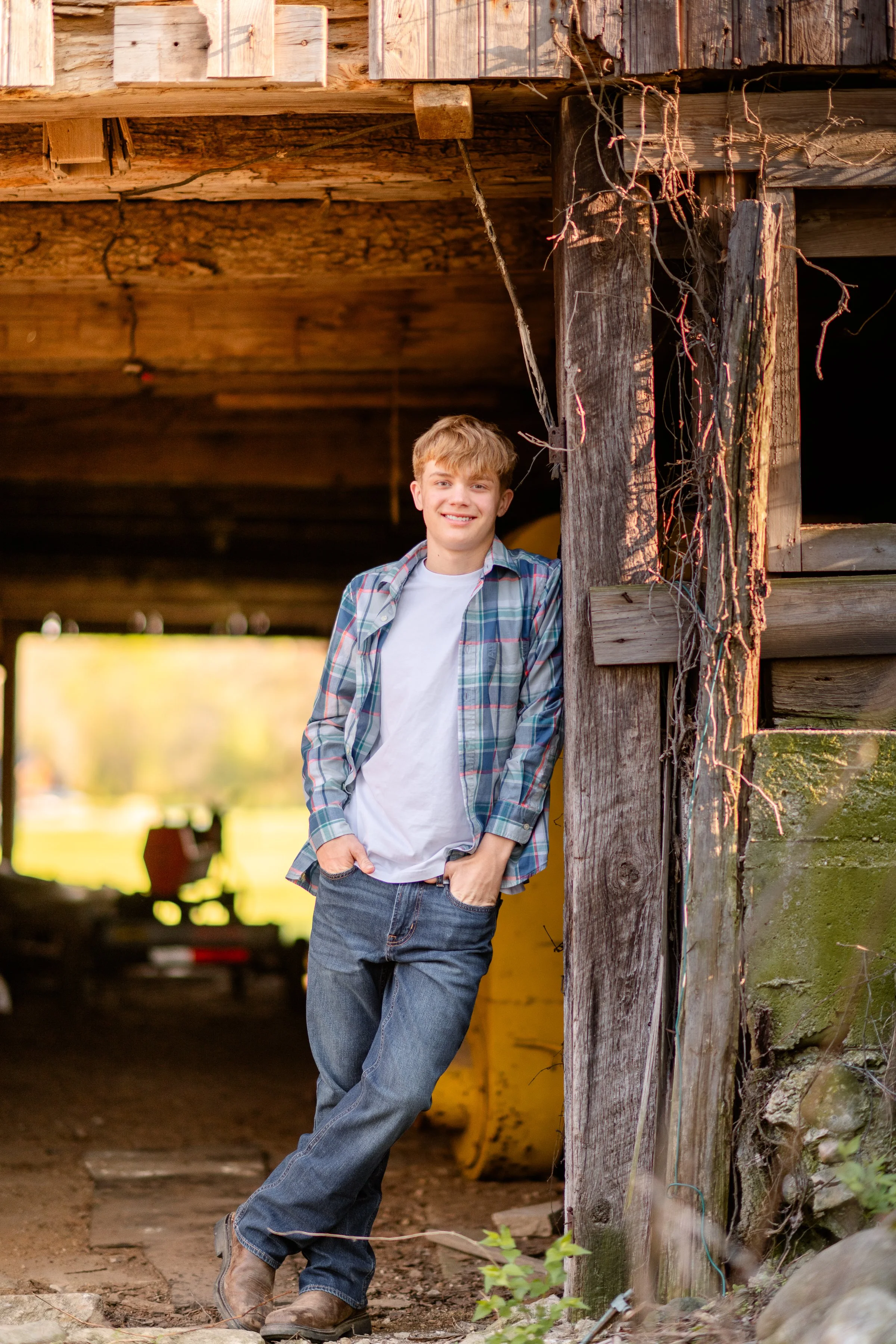 A young man with blonde hair and a friendly smile stands outdoors leaning against a wooden support post under a weathered wooden bridge, wearing a plaid shirt over a white t-shirt, jeans, and boots, with a natural background of green and yellow foliage.