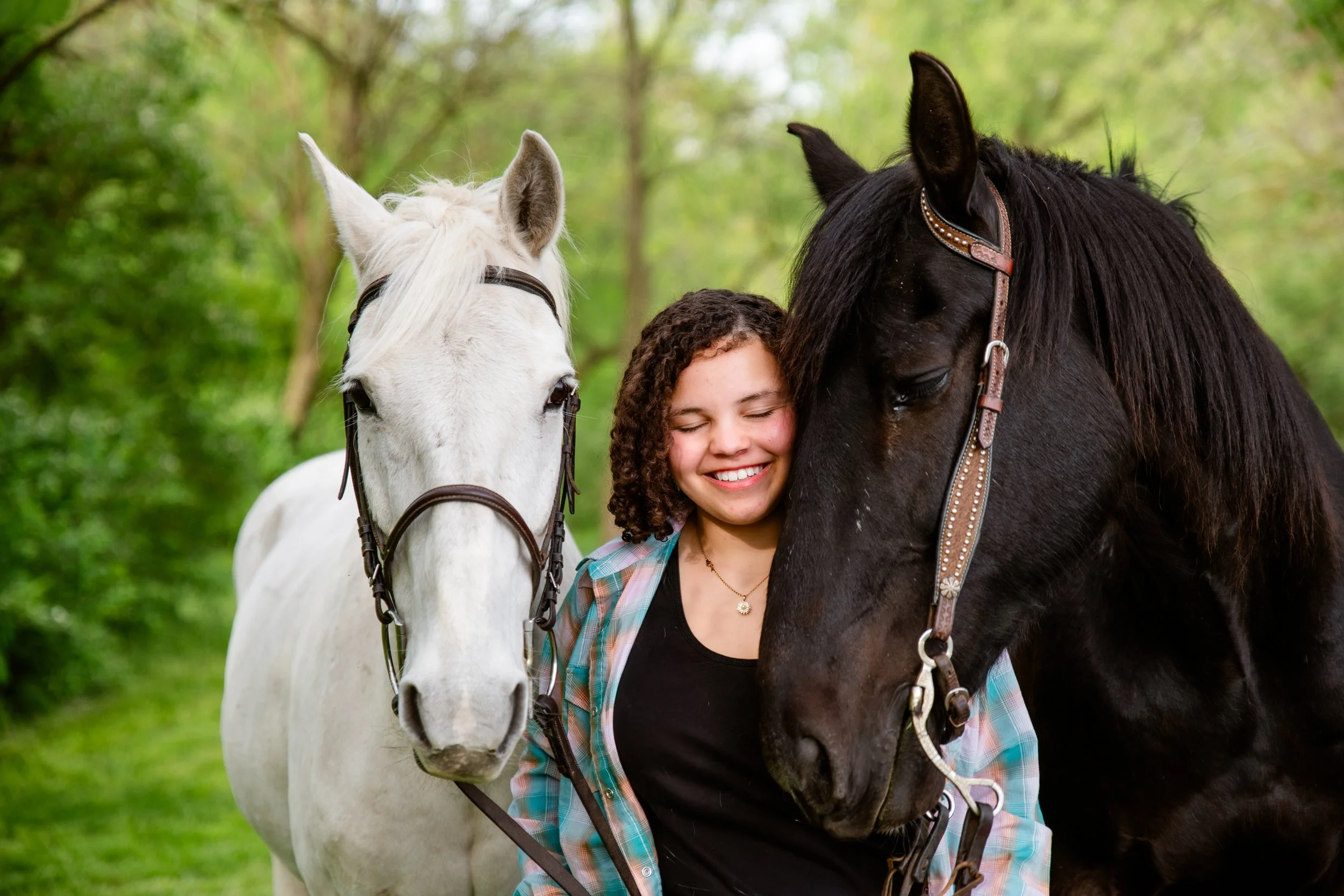 A girl with curly hair smiling with eyes closed, hugging a black horse, and standing next to a white horse, in a green outdoor setting.