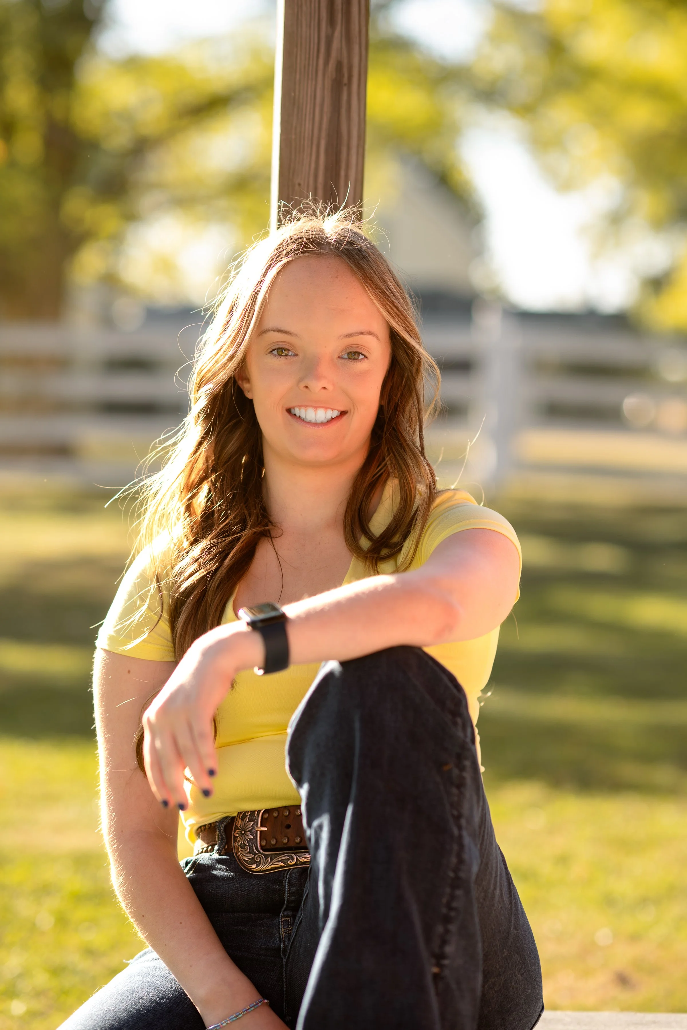 A young woman with long red hair, wearing a yellow shirt and black jeans, smiling and sitting outdoors in a park with green trees in the background. She is wearing a watch and bracelet, and holding a black jacket, with bright sunlight highlighting her face and hair.