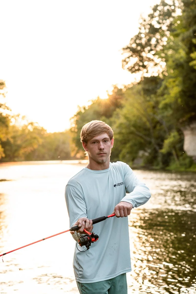 Young man fishing at a river during sunset, holding a fishing rod and looking at the camera, surrounded by greenery.