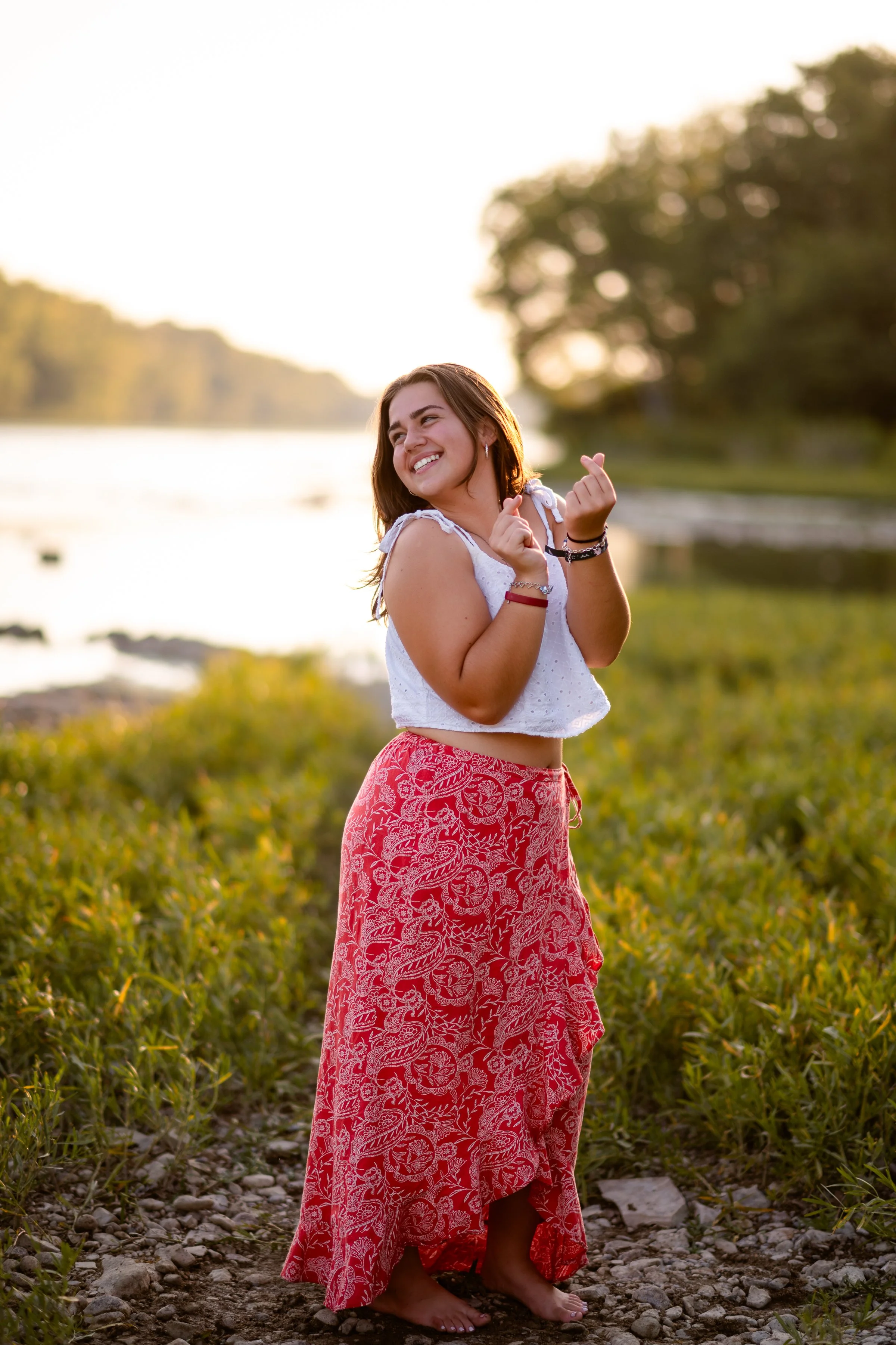 A woman standing barefoot outdoors on a rocky path near a body of water, wearing a white sleeveless top and a long red skirt with white floral patterns, smiling and looking to her left at sunset.