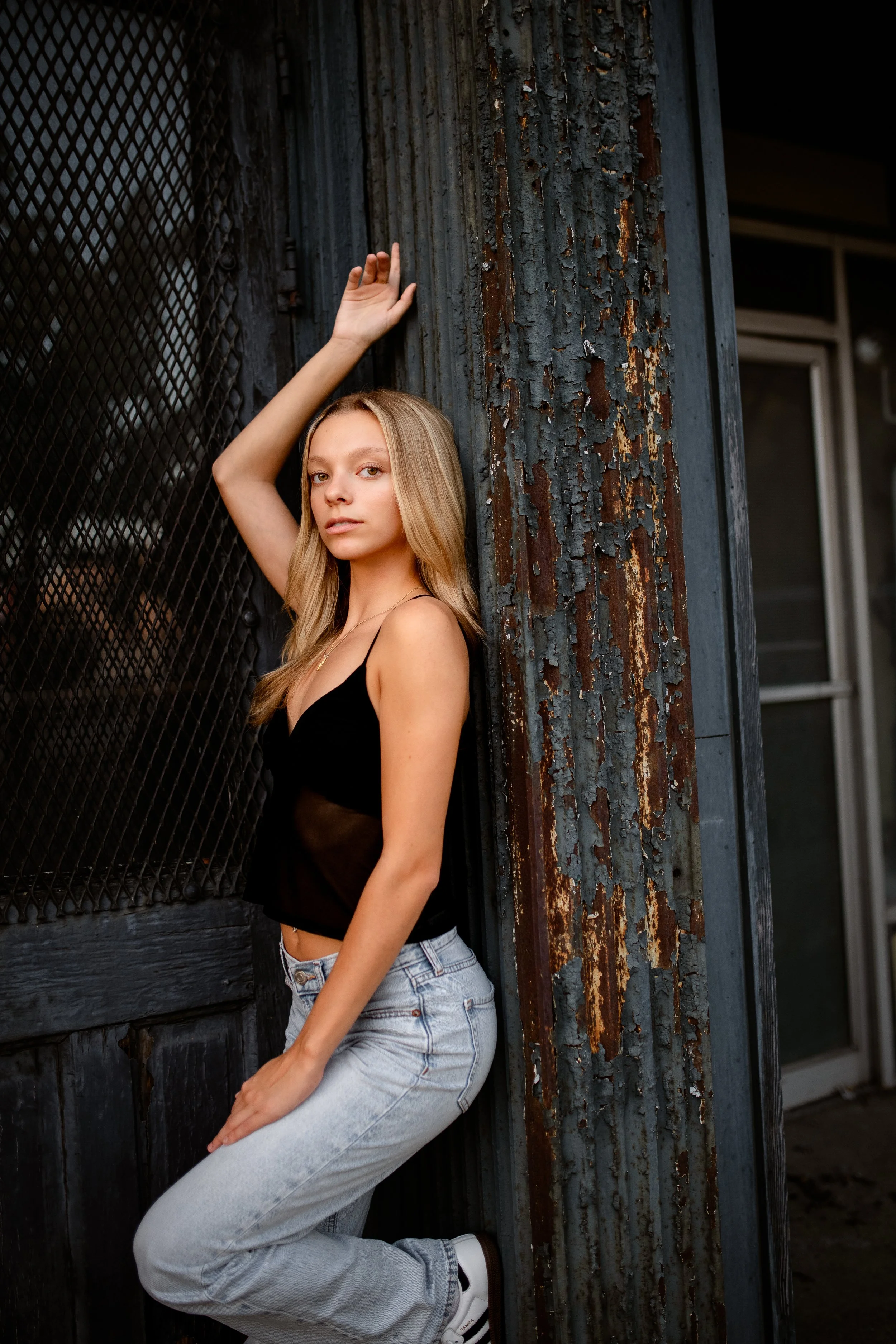 A young woman with blonde hair wearing a black tank top and light blue jeans is leaning against a weathered, rusty metal wall with peeling paint. She has her right arm raised with her hand near her head, looking at the camera with a neutral expression.