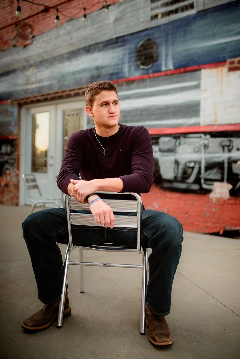 A young man sitting on a metal chair outdoors, wearing a black long-sleeve shirt, dark jeans, and brown boots, with a cross necklace, in front of a rustic wall with painted train artwork.