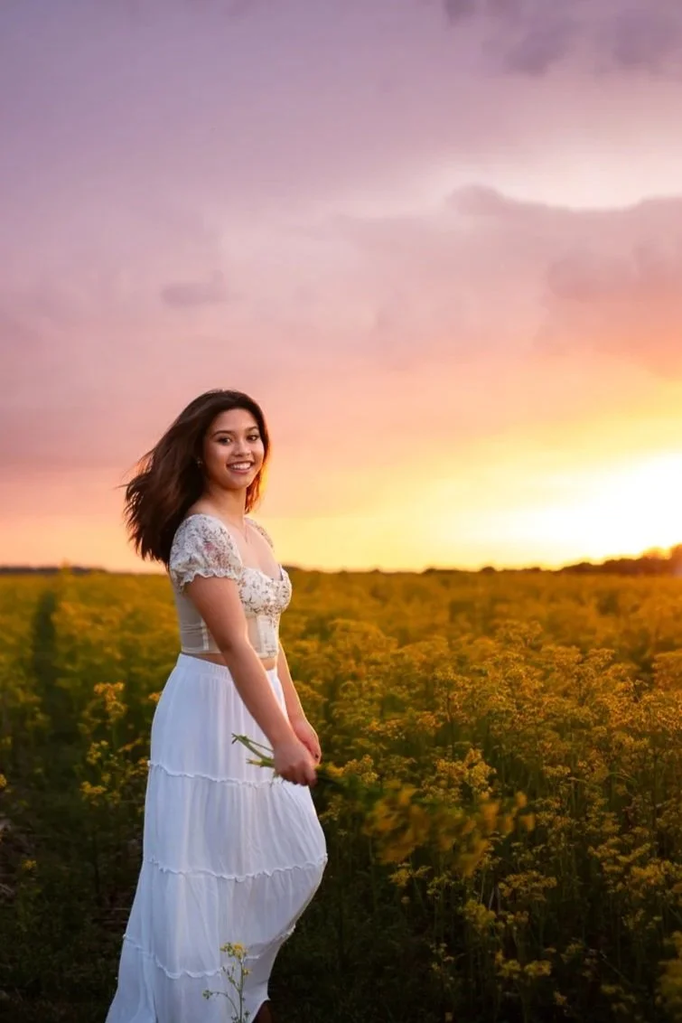 A young woman in a white summer dress standing in a field of yellow flowers at sunset