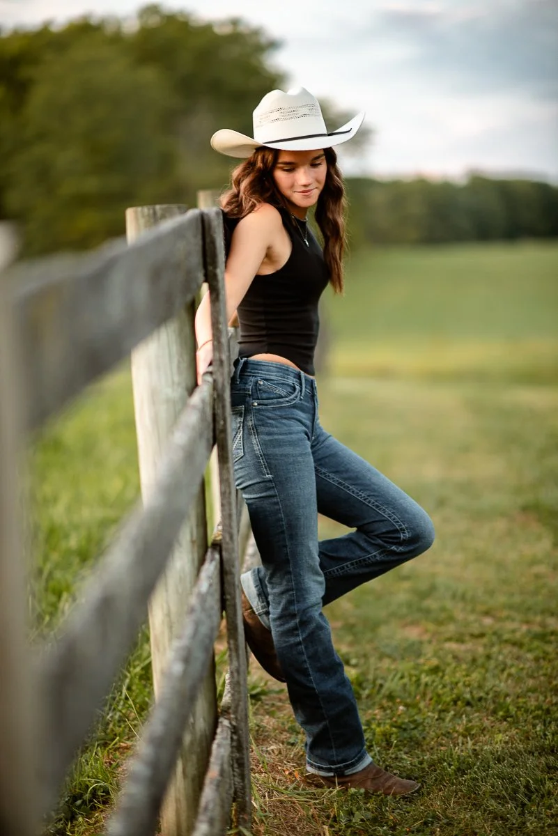 A young woman with long brown hair wearing a white cowboy hat, black sleeveless top, and jeans leaning against a wooden fence in a grassy outdoor area with trees in the background.