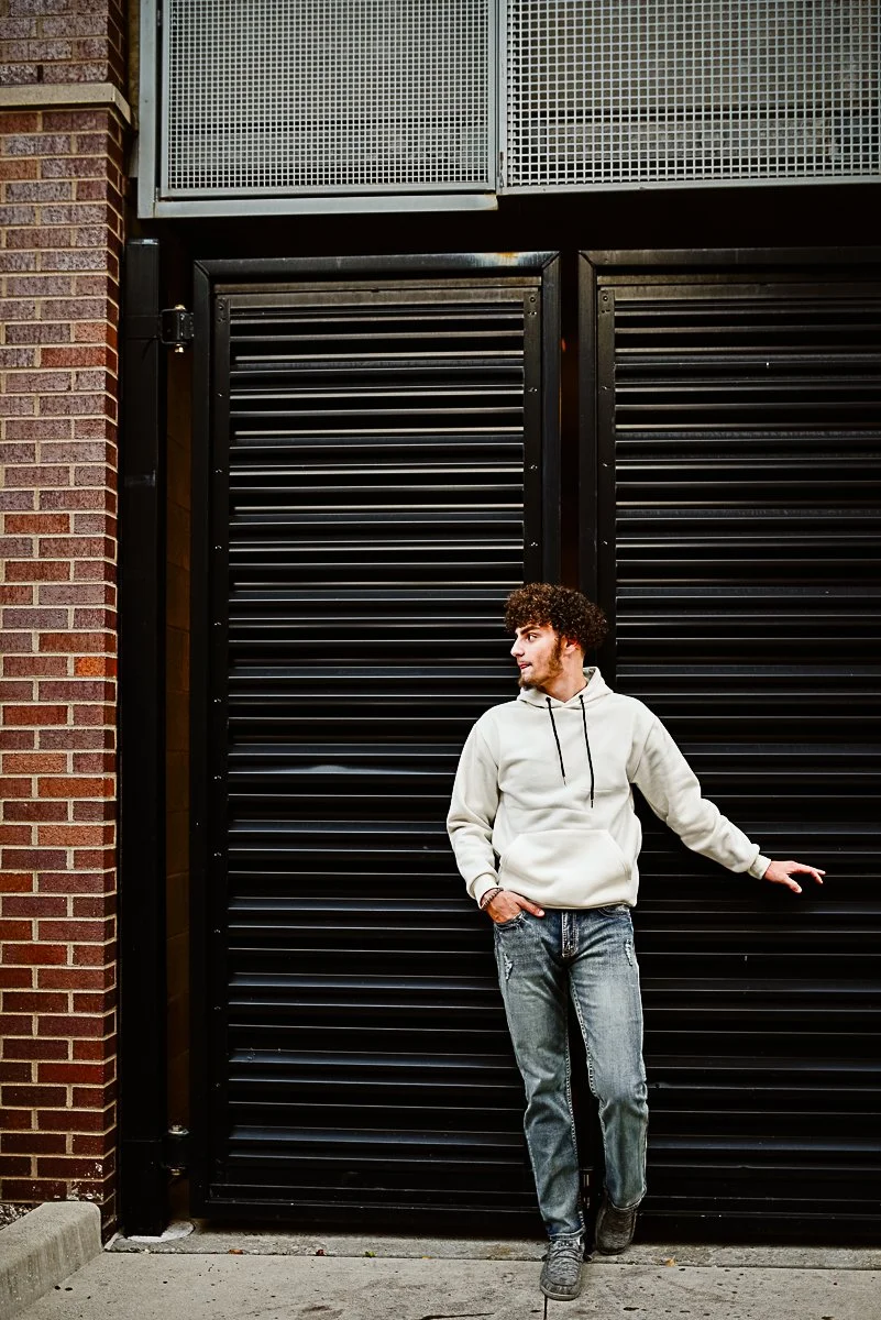 A young man with curly hair wearing a white hoodie and distressed jeans standing outside in front of a large black industrial gate with brick wall on the left side.