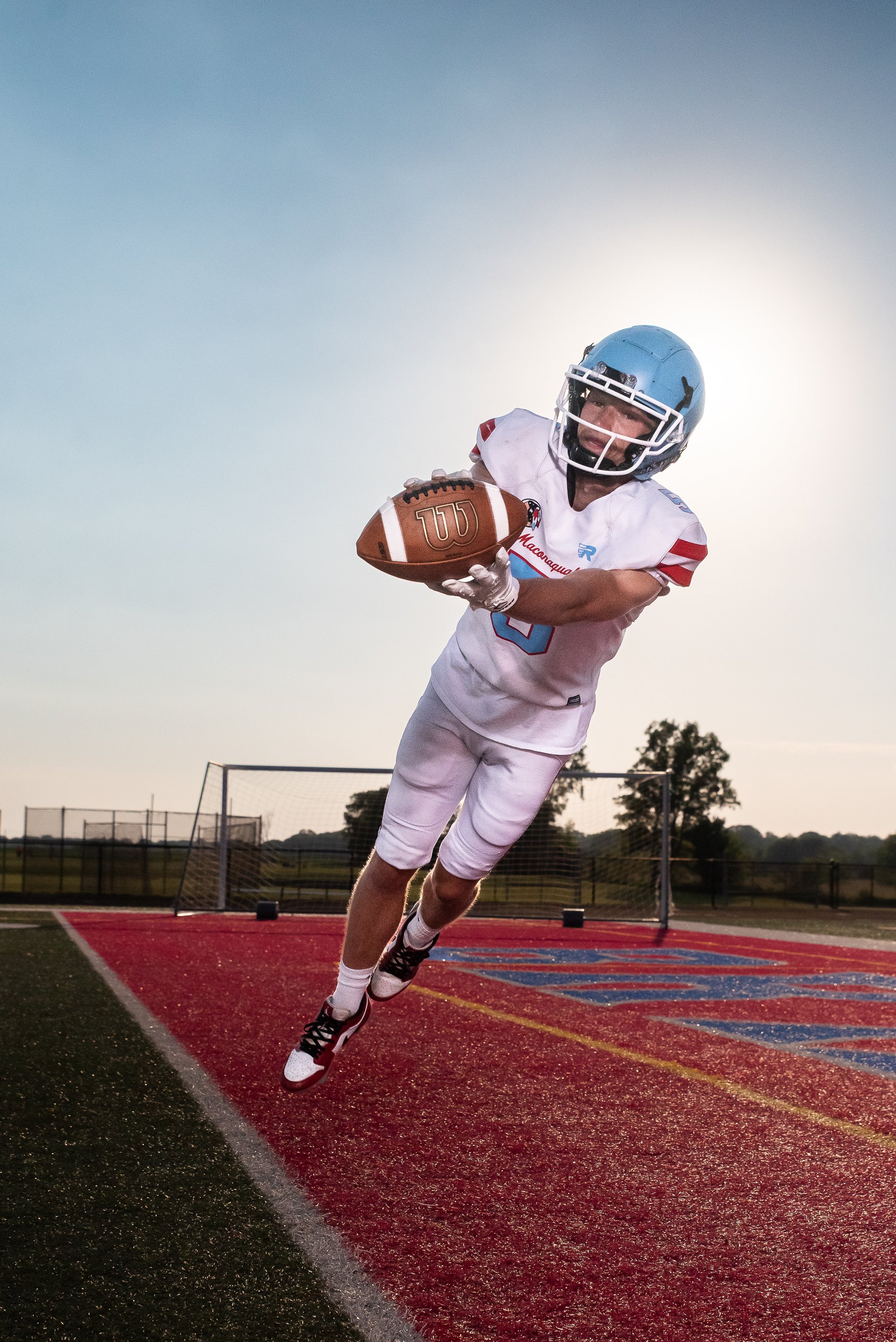 A football player in a white uniform and helmet is jumping and reaching for a football on a red and black sports field during sunset.