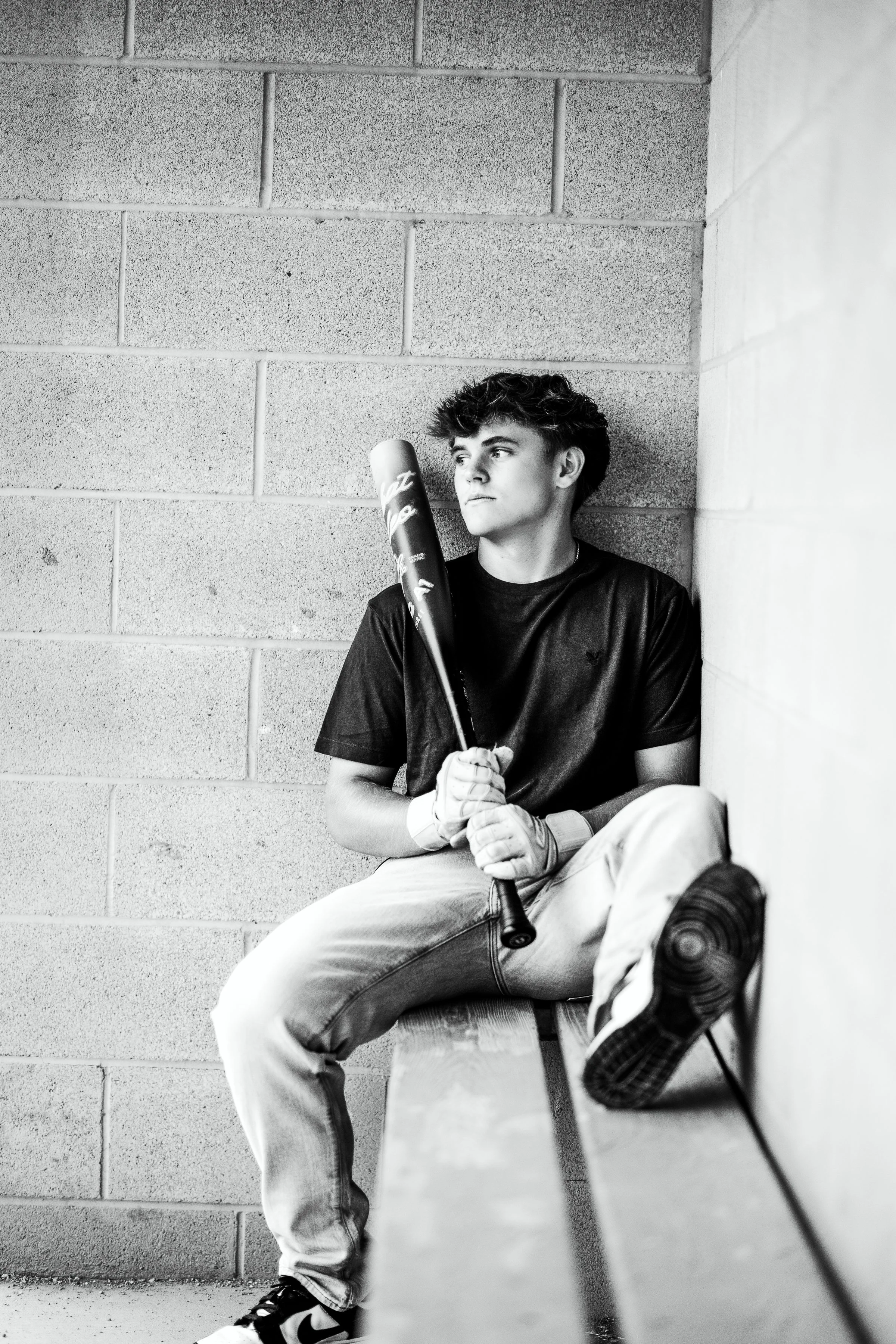 A black and white photo of a young man sitting on a stool against a brick wall, holding a baseball bat, wearing a t-shirt, jeans, sneakers, and baseball gloves, with one leg crossed over the other.