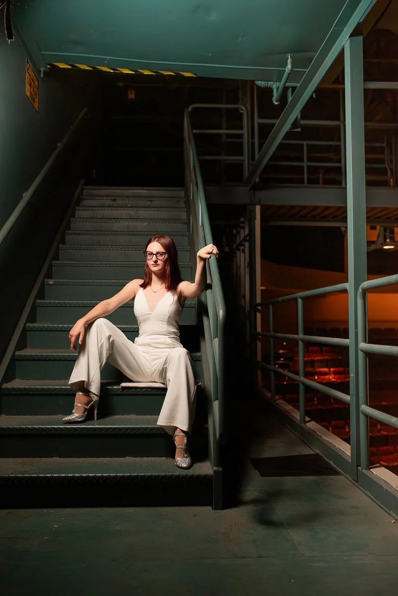 A woman with glasses in a white jumpsuit and silver heels sitting on a metal staircase in an industrial or parking garage-like setting.
