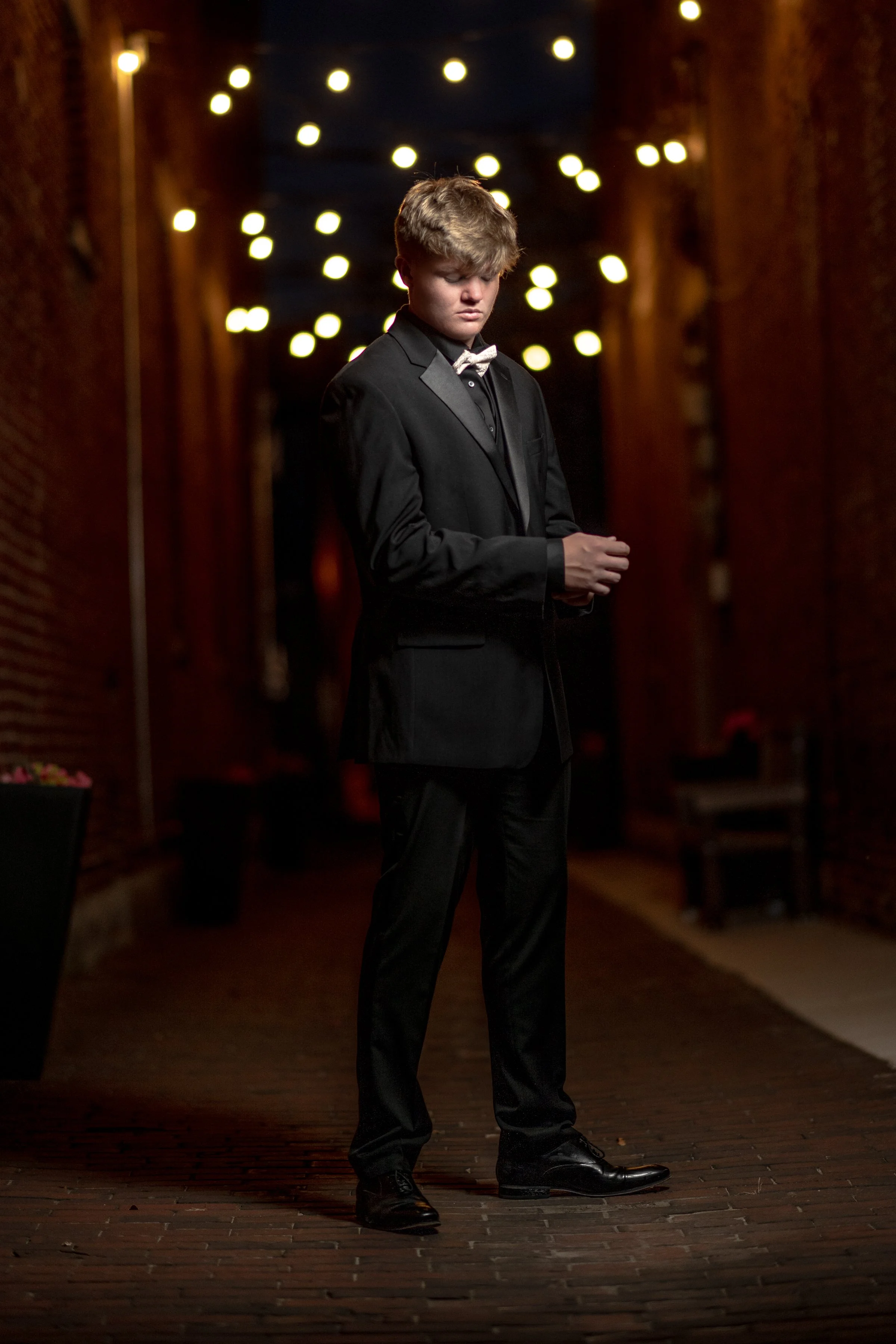 A young man in a black suit with a grey bow tie standing in a dimly lit outdoor area with string lights overhead, with a serious expression and hands clasped.