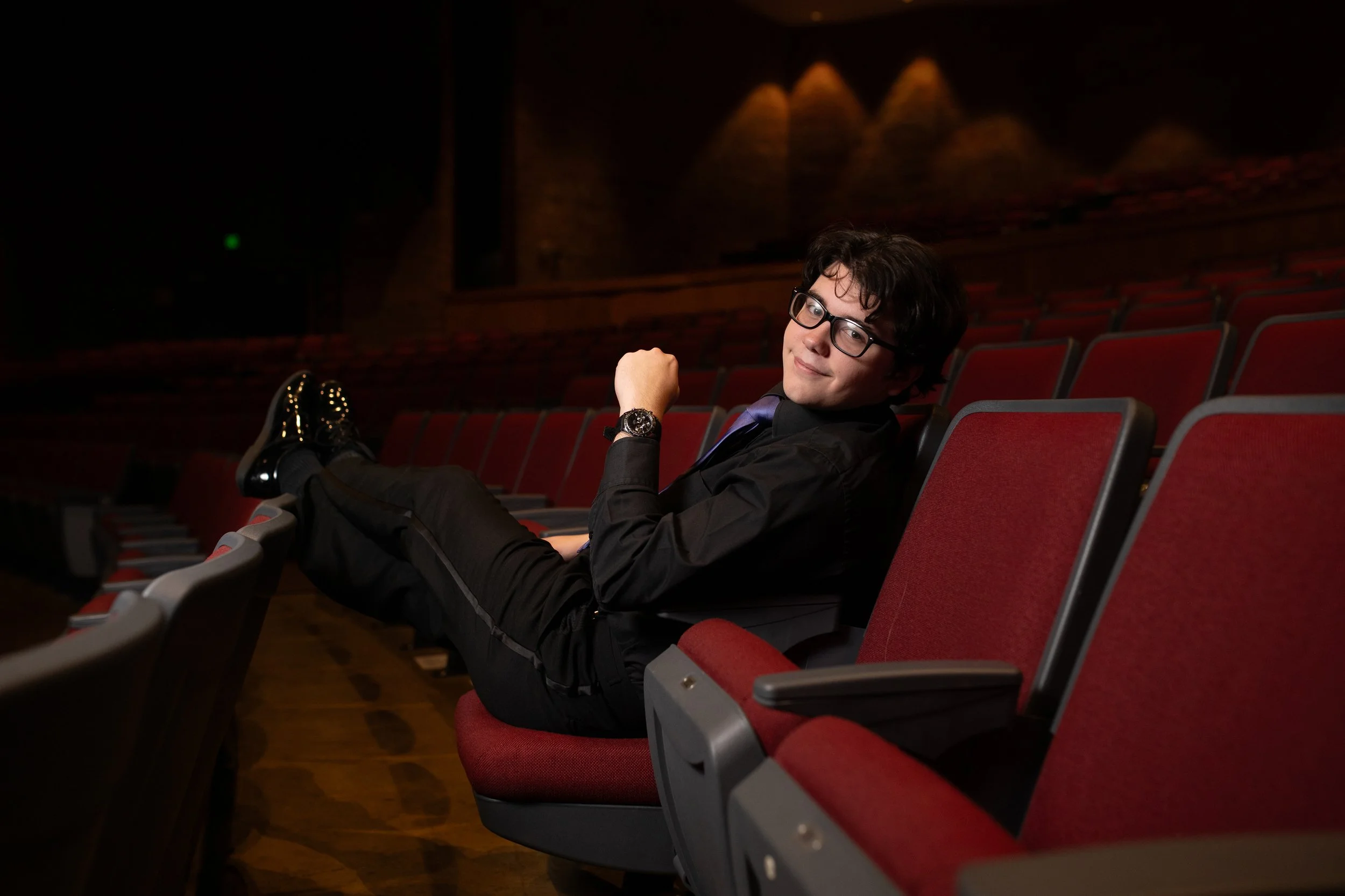 Young man with glasses and a watch reclining in a theater seat with red upholstery, smiling, with empty theater seats and dim lighting in the background.