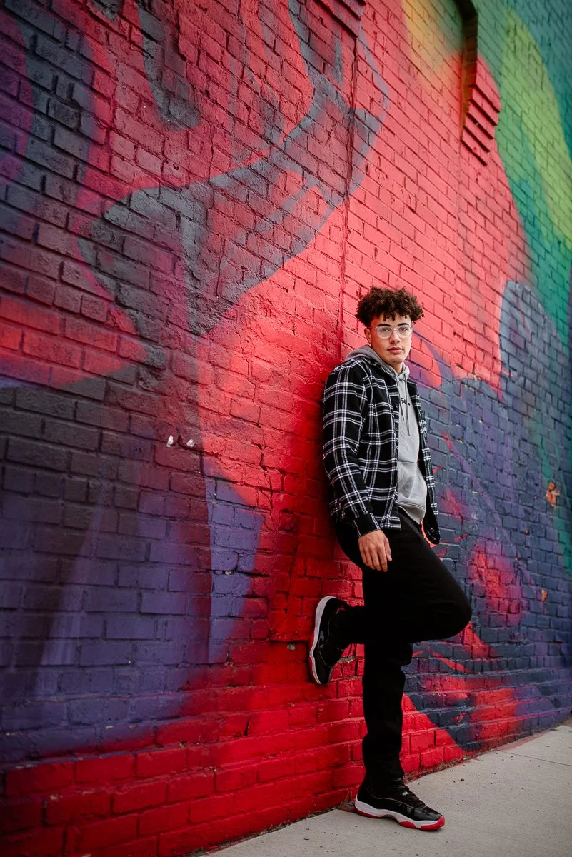 A young man with curly hair and glasses posing against a colorful graffiti mural on a brick wall, wearing a black and white checkered hoodie, gray hoodie, black pants, and black sneakers.