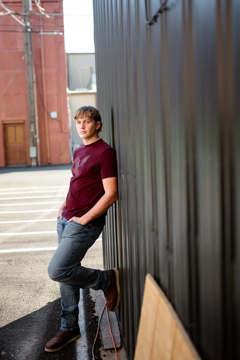 Young man leaning against a metal wall in an urban alleyway, with one foot resting on a curb and casual clothing.
