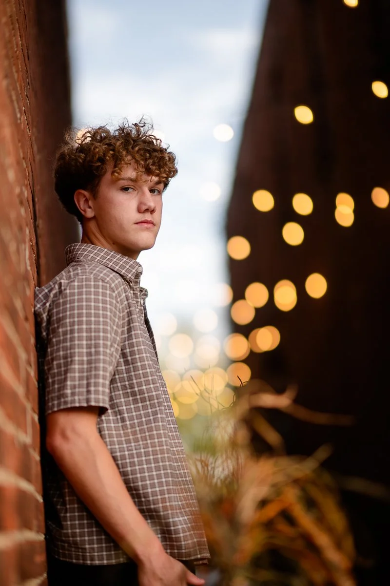 A young man with curly hair in a checkered shirt leaning against a brick wall during dusk with string lights in the background.