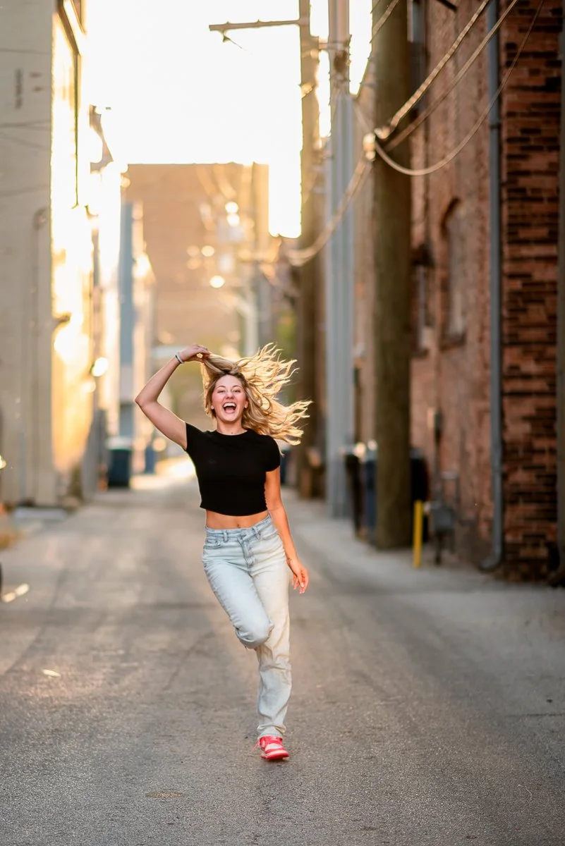 Young woman running joyfully down an alleyway at sunset, hair flowing, wearing a black crop top, light jeans, and red sneakers.