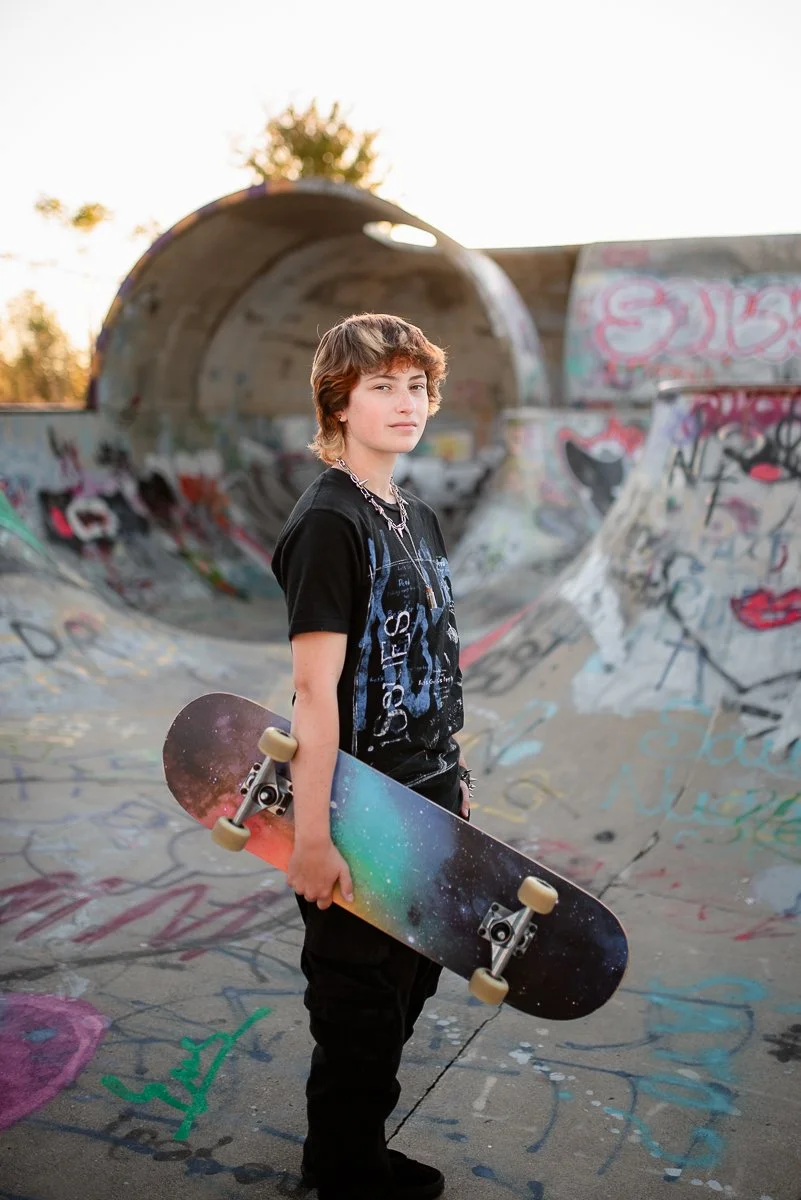 A young person with short curly hair holding a skateboard at a graffiti-covered skate park during sunset.