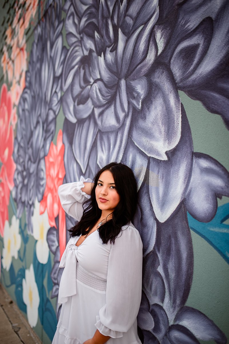 A young woman with dark hair wearing a white dress, standing against a colorful floral mural wall.