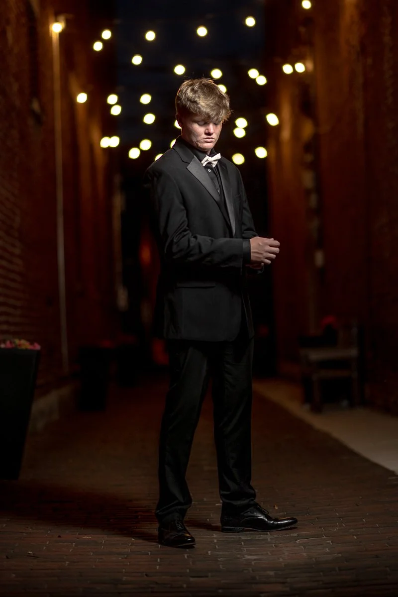 A young man in a tuxedo standing in a dimly lit alley at night with string lights overhead, looking down with a serious expression.
