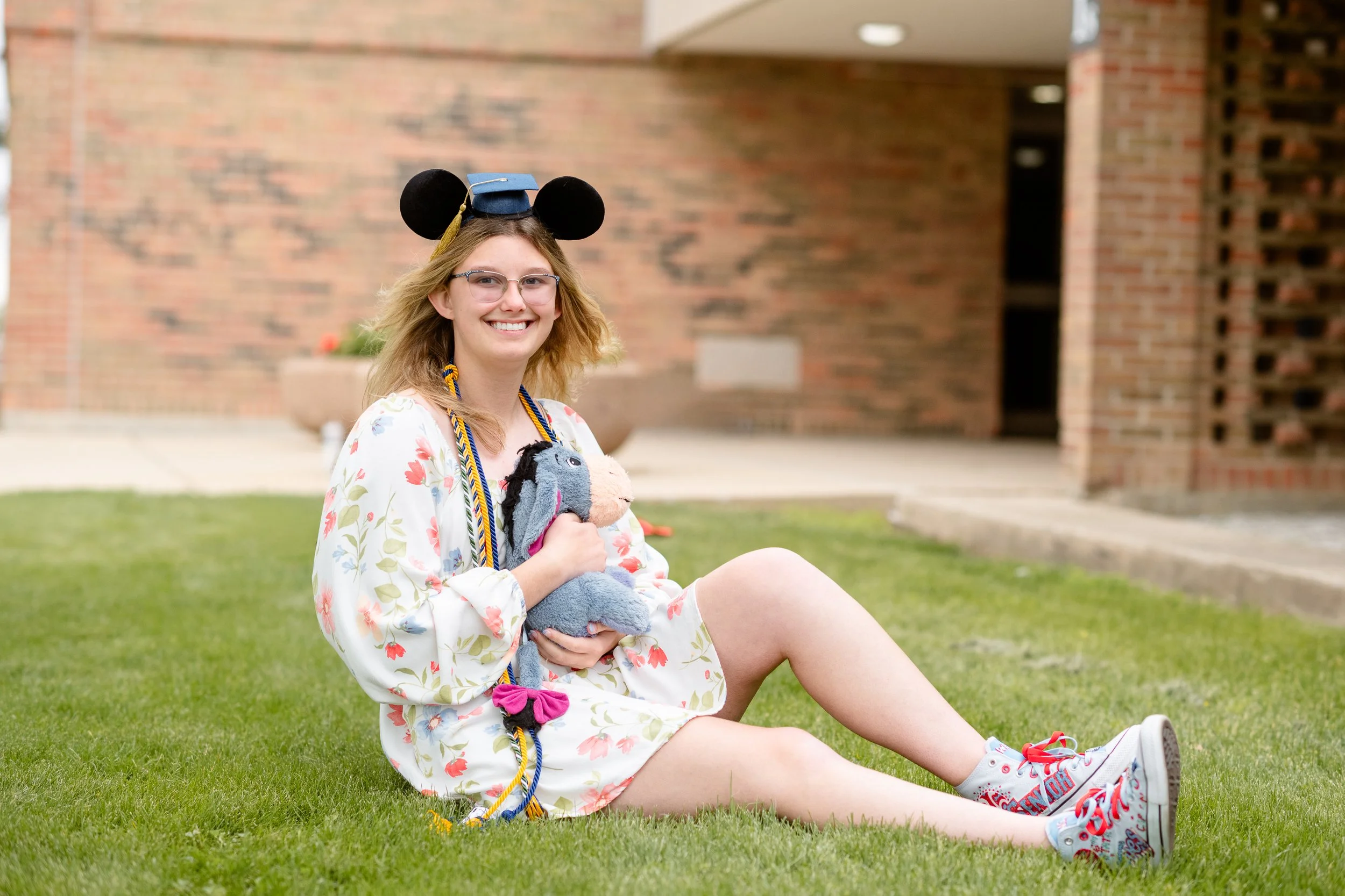 A young woman with glasses, blonde hair, and a big smile, sitting on a grassy area outside a building with brick walls. She is wearing a floral dress, a graduation cap, and Mickey Mouse ears, and holding a stuffed animal. Her shoes are white sneakers with colorful designs.