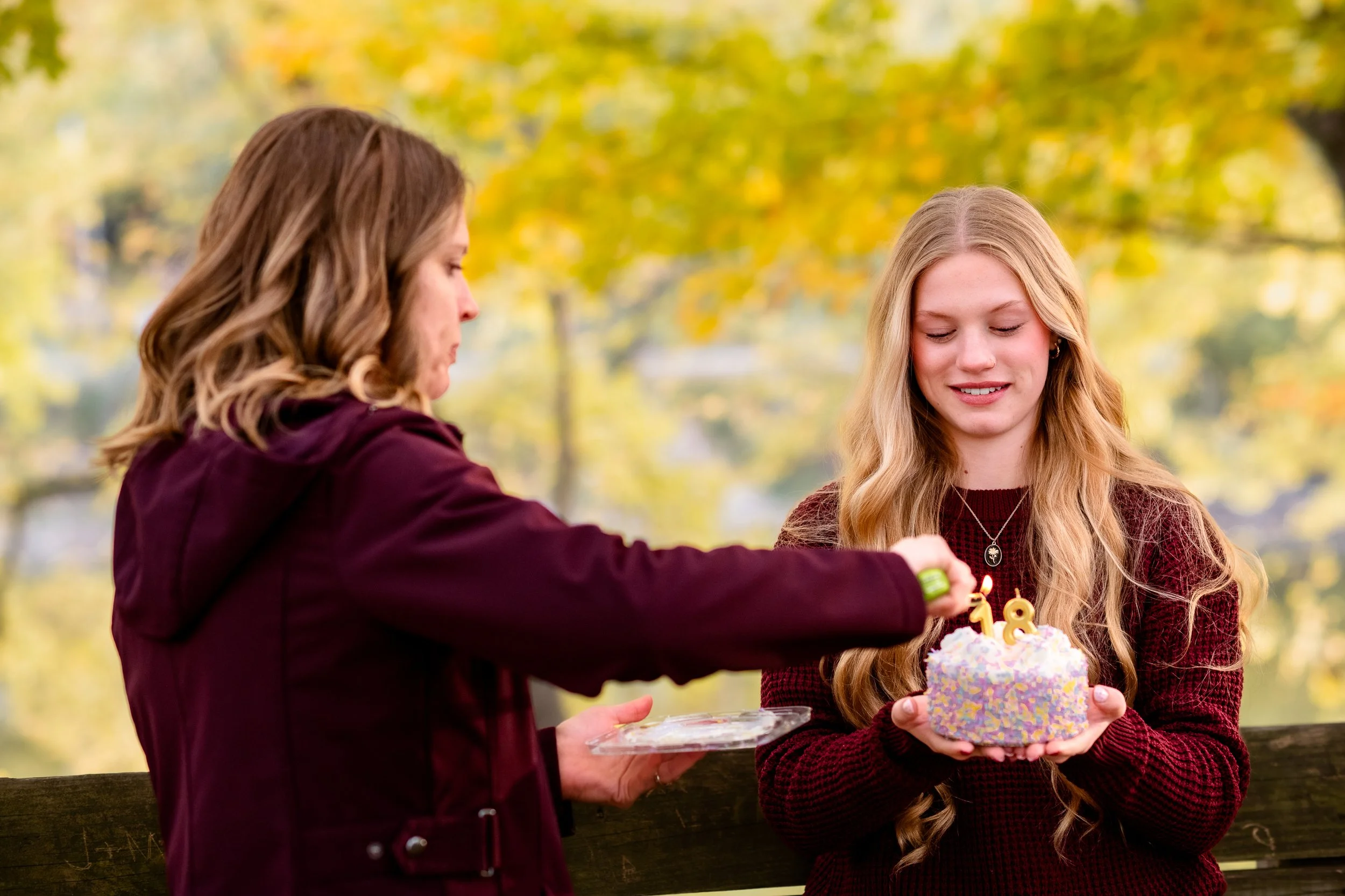 Two women celebrating a birthday outdoors, with one woman holding a colorful birthday cake with lit candles. The other woman is applying something on her wrist, possibly a bracelet or watch. The background shows autumn trees with yellow leaves.
